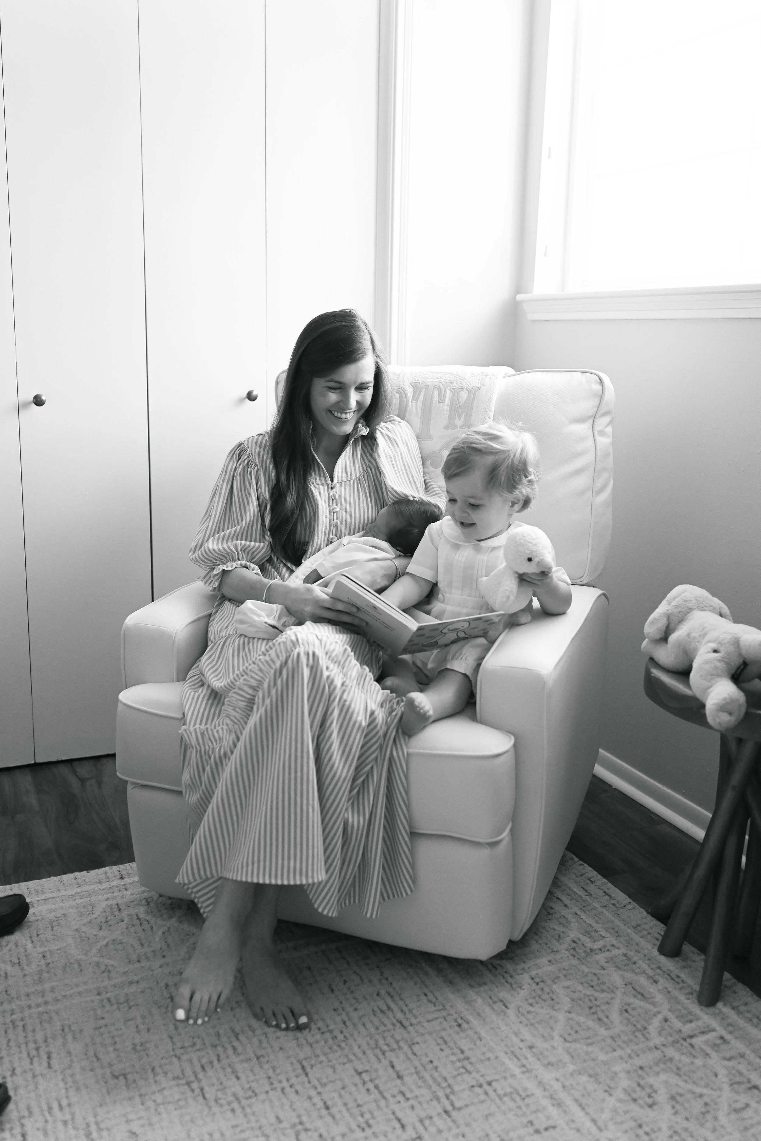 A woman sitting in a chair with two young children, one breastfeeding and the other holding a stuffed duck, in a cozy living room with bright windows.