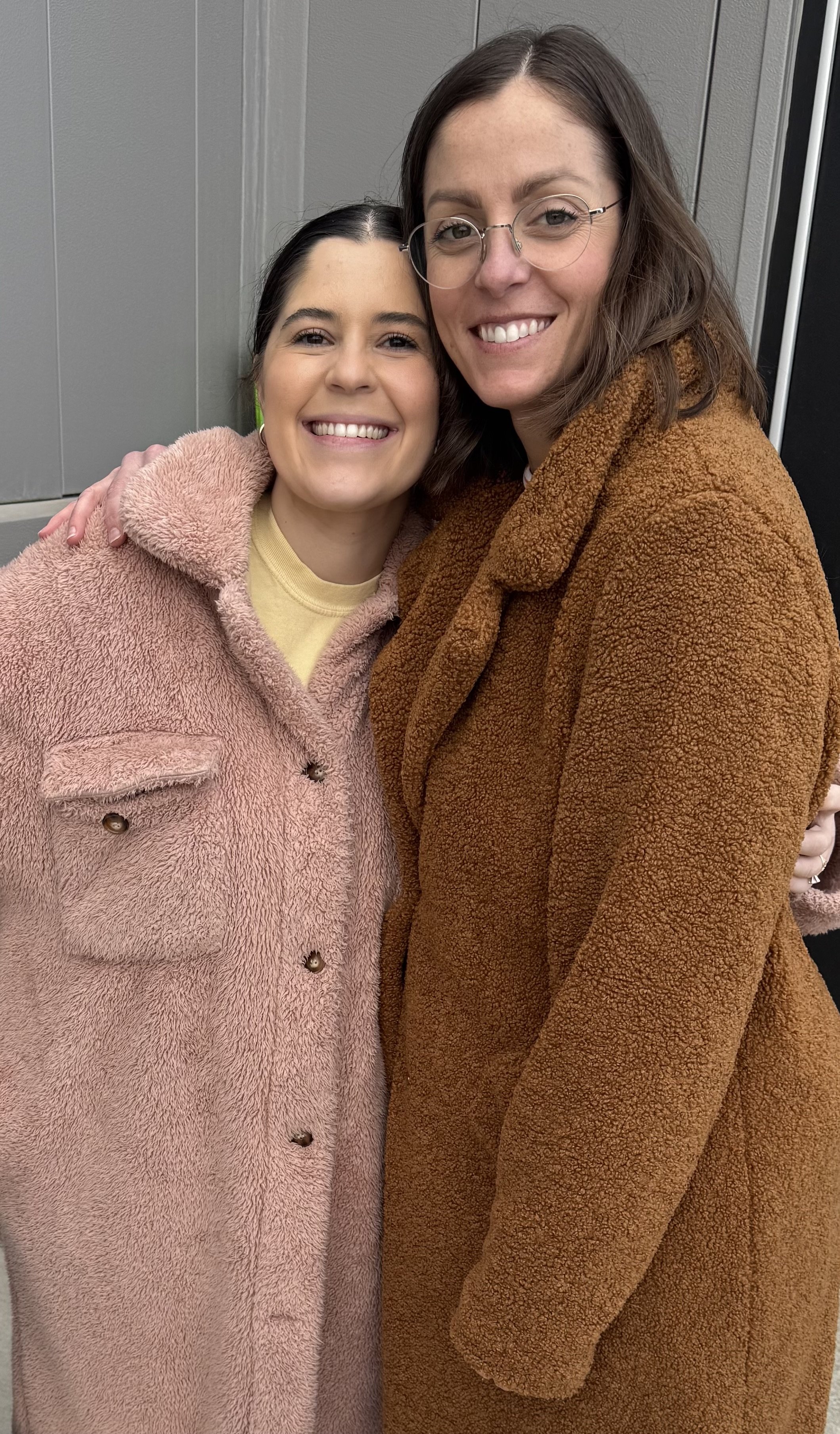 Two smiling women in warm coats, one in pink and the other in brown, standing close together with their arms around each other, inside a grey-paneled room.