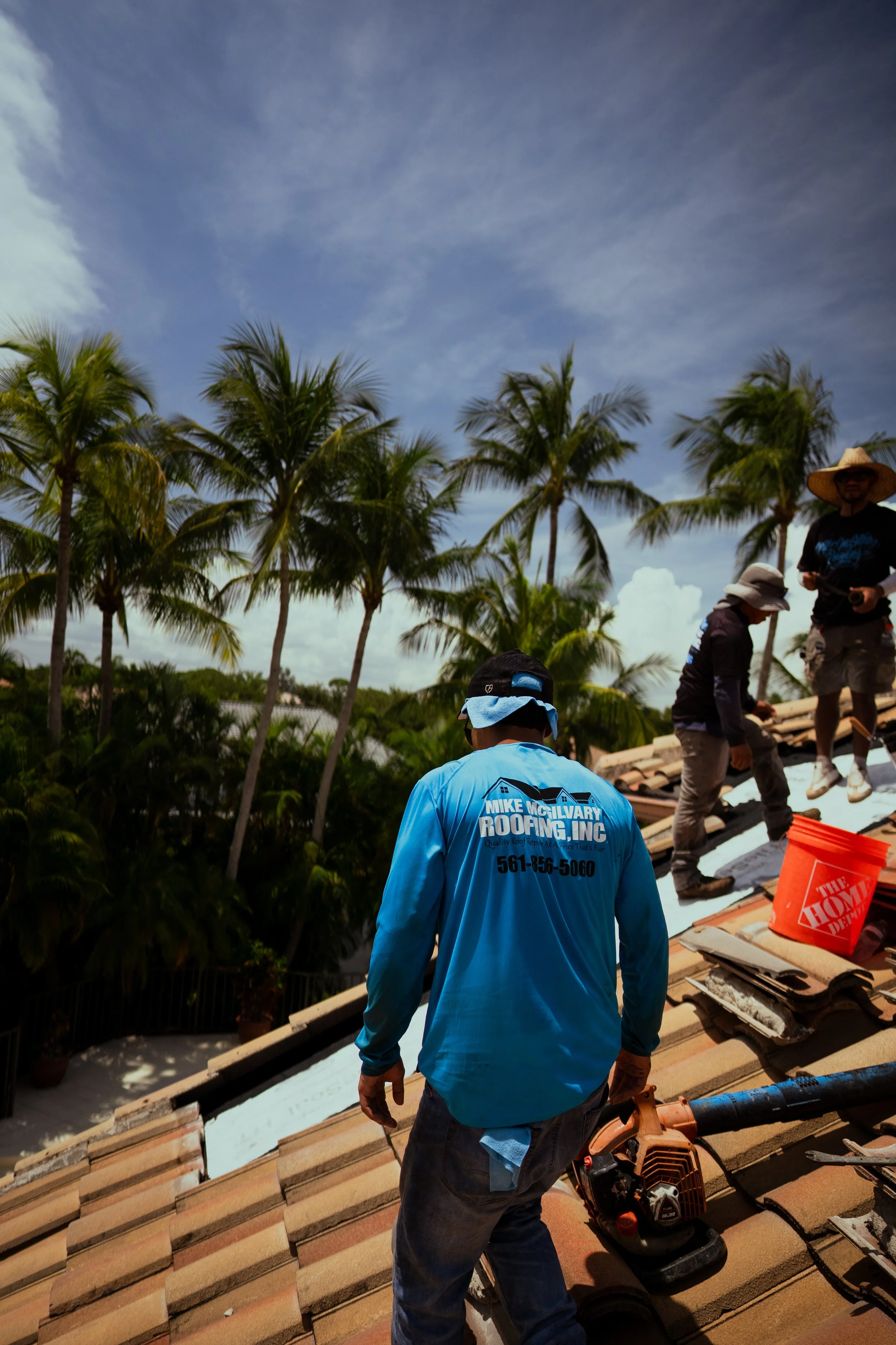 Roofing workers installing tiles on a rooftop under a blue sky with palm trees in the background.