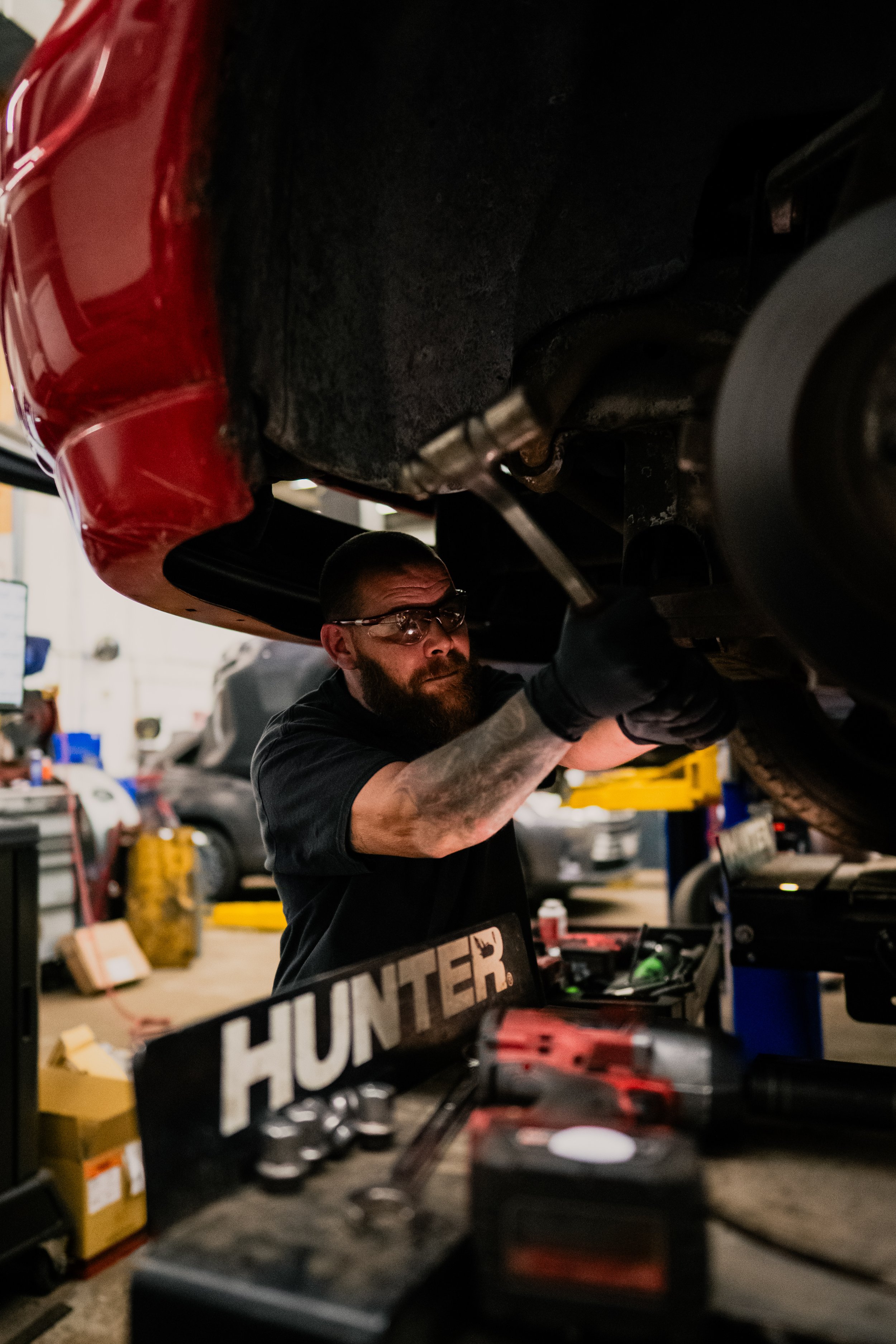A mechanic working underneath a car in a garage, wearing safety glasses and black gloves, with tools and equipment around.