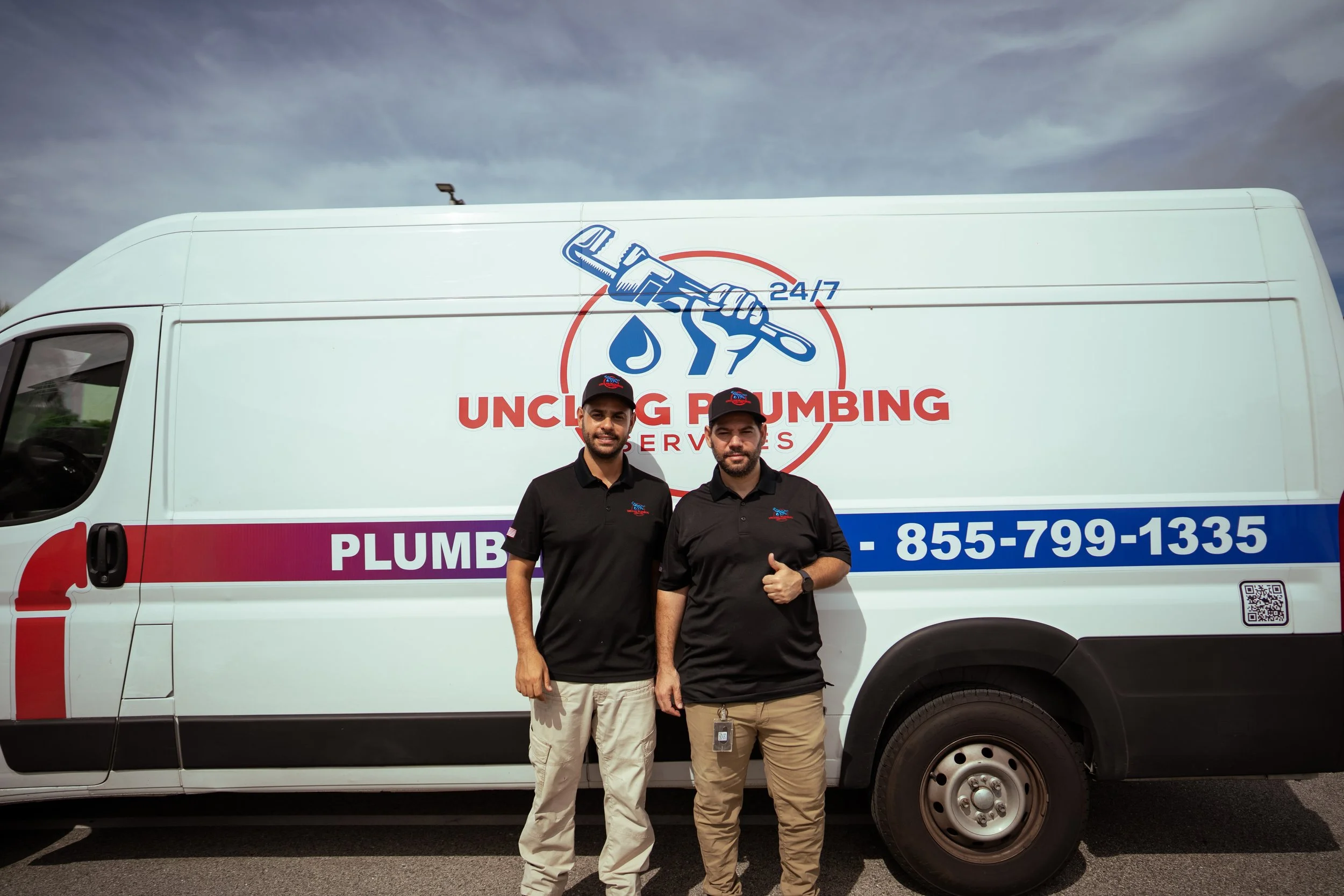 Two technicians in black uniforms standing in front of a white plumbing service van with phone number 855-799-1335, company logo, and a graphic of a wrench and water drop.