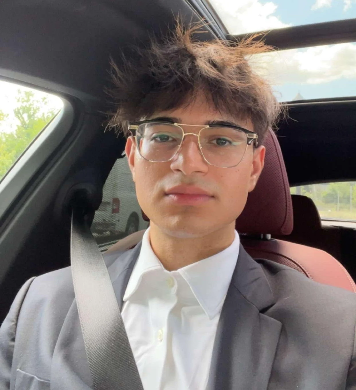 A young man with brown hair wearing glasses, a white shirt, and a suit jacket sitting in a car, taking a selfie.