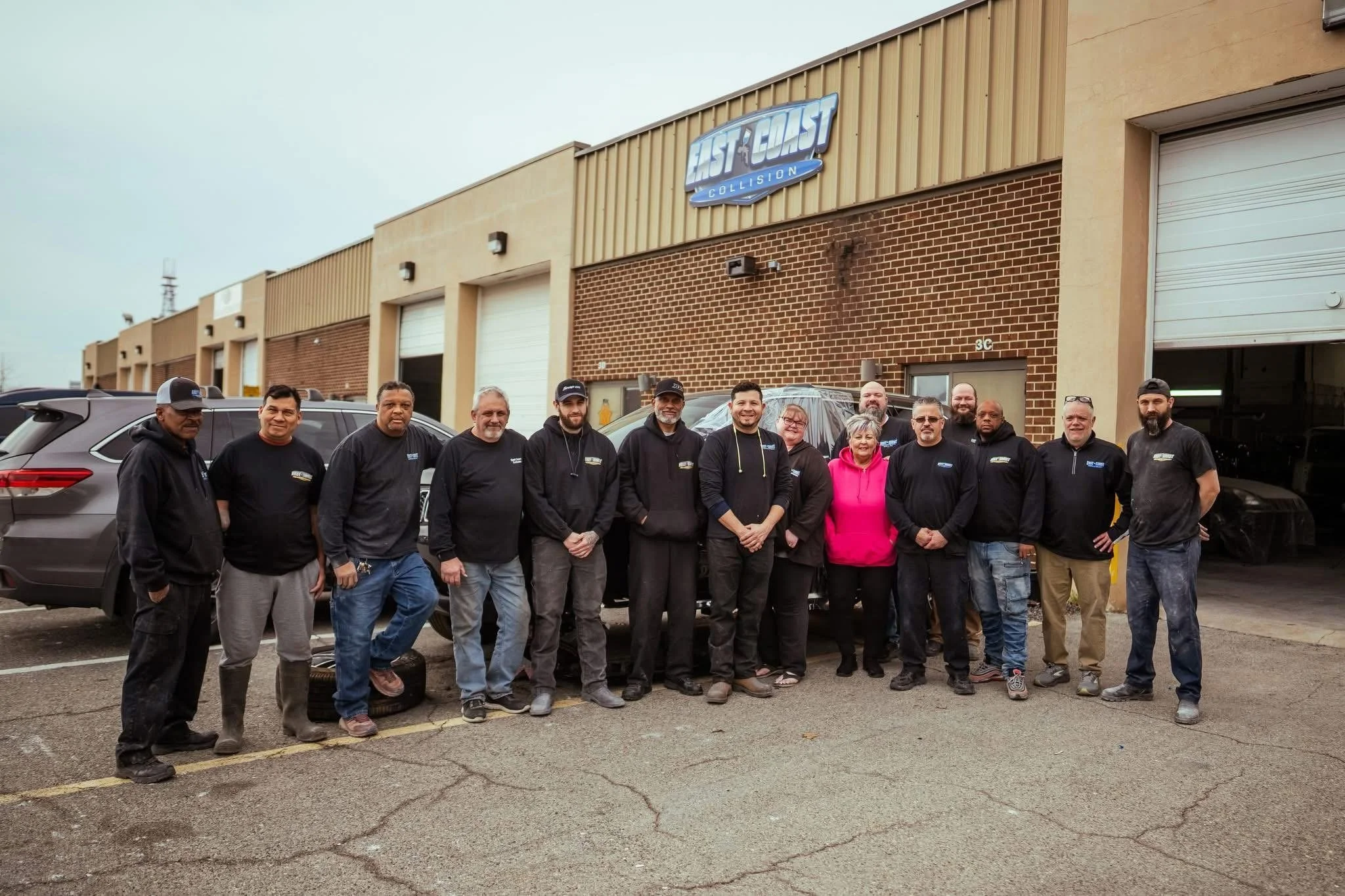 Group of people standing outside an auto repair shop called 'East Coast Collision,' some wearing black hoodies, one person in pink, standing in front of several parked cars.
