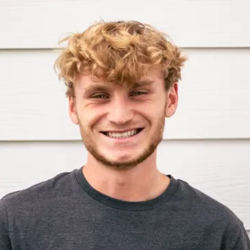 A young man with curly blonde hair and a beard smiling, wearing a dark gray t-shirt, standing in front of a white wooden wall.