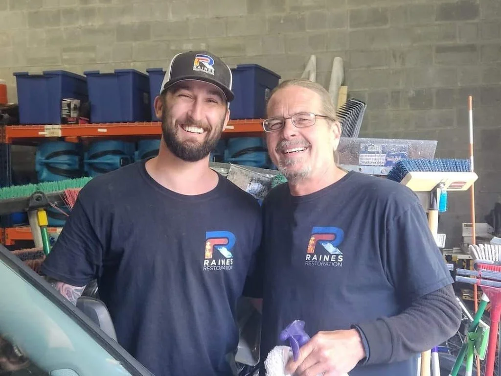 Two men wearing black Raines Restoration t-shirts smiling and standing close together inside a warehouse or workshop, with various tools and equipment in the background.