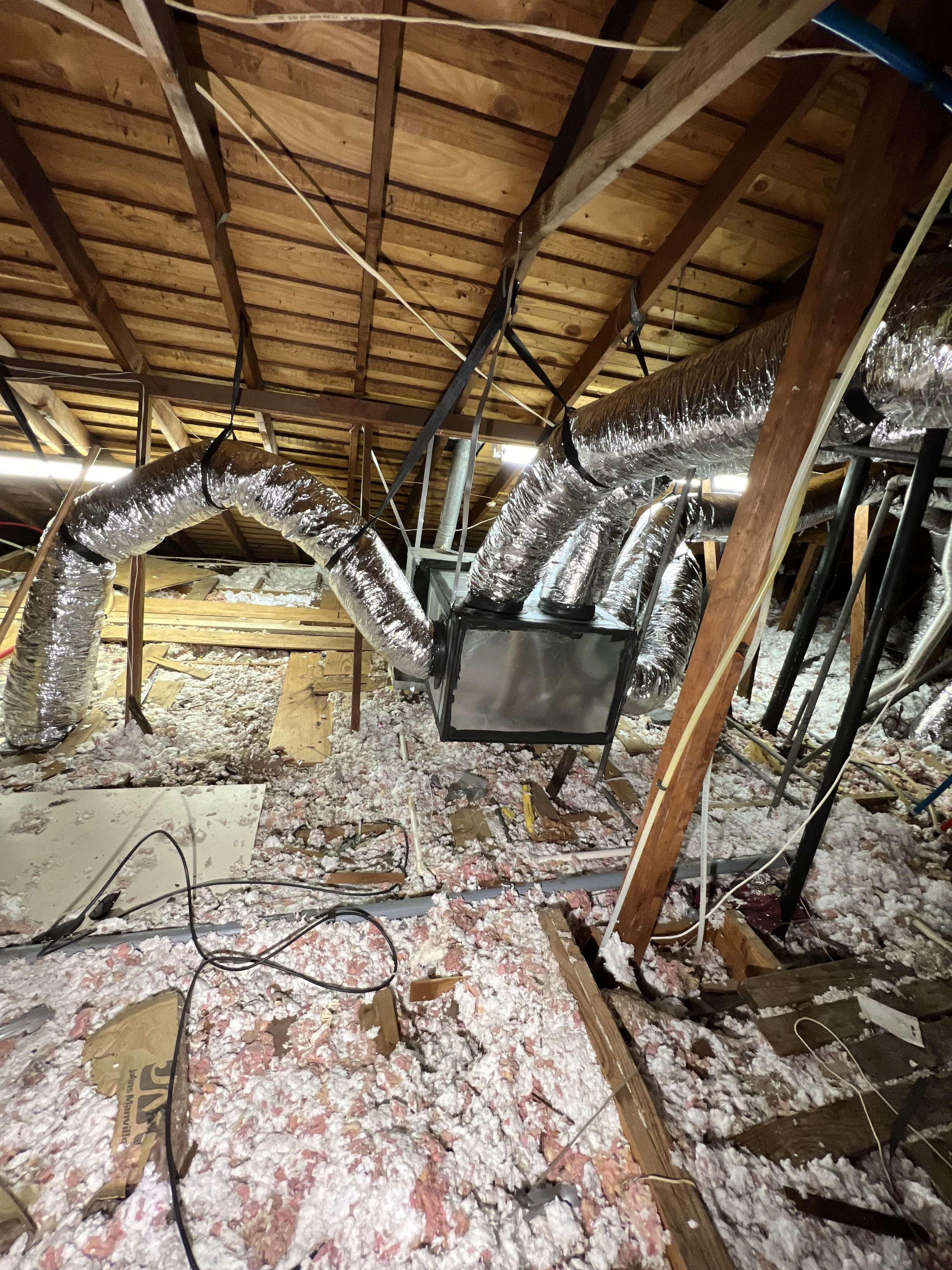 Enclosed attic space with wooden rafters, foil ductwork, electrical wires, and insulation scattered across the floor.