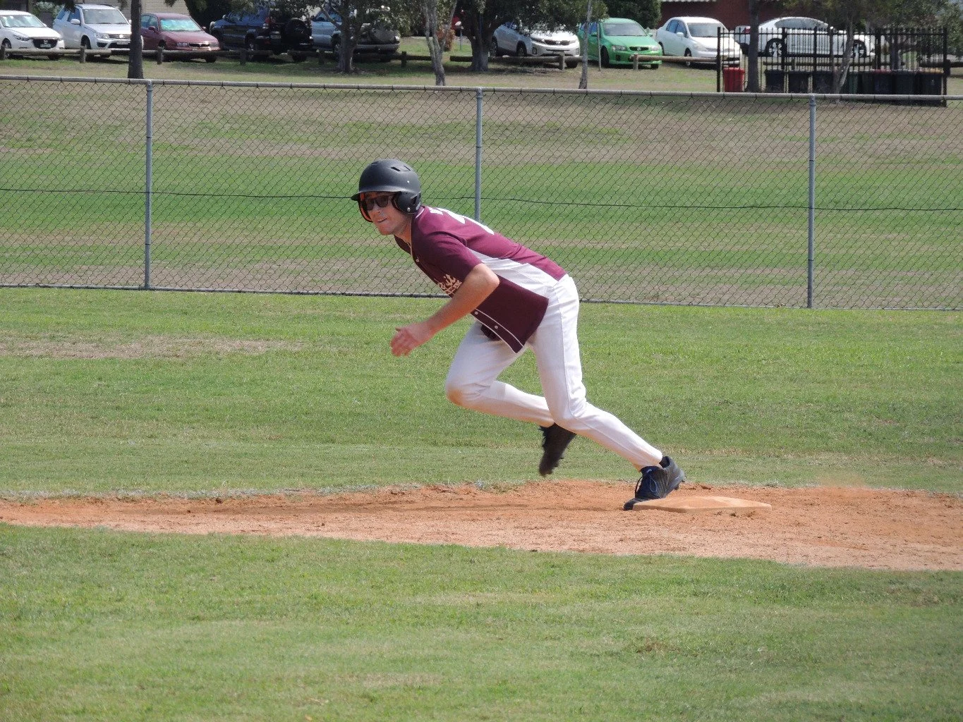 A baseball player wearing sunglasses, a helmet, a maroon and white jersey, and white pants, is crouching and ready to run on a grassy field near a dirt base, with parked cars and a chain-link fence in the background.