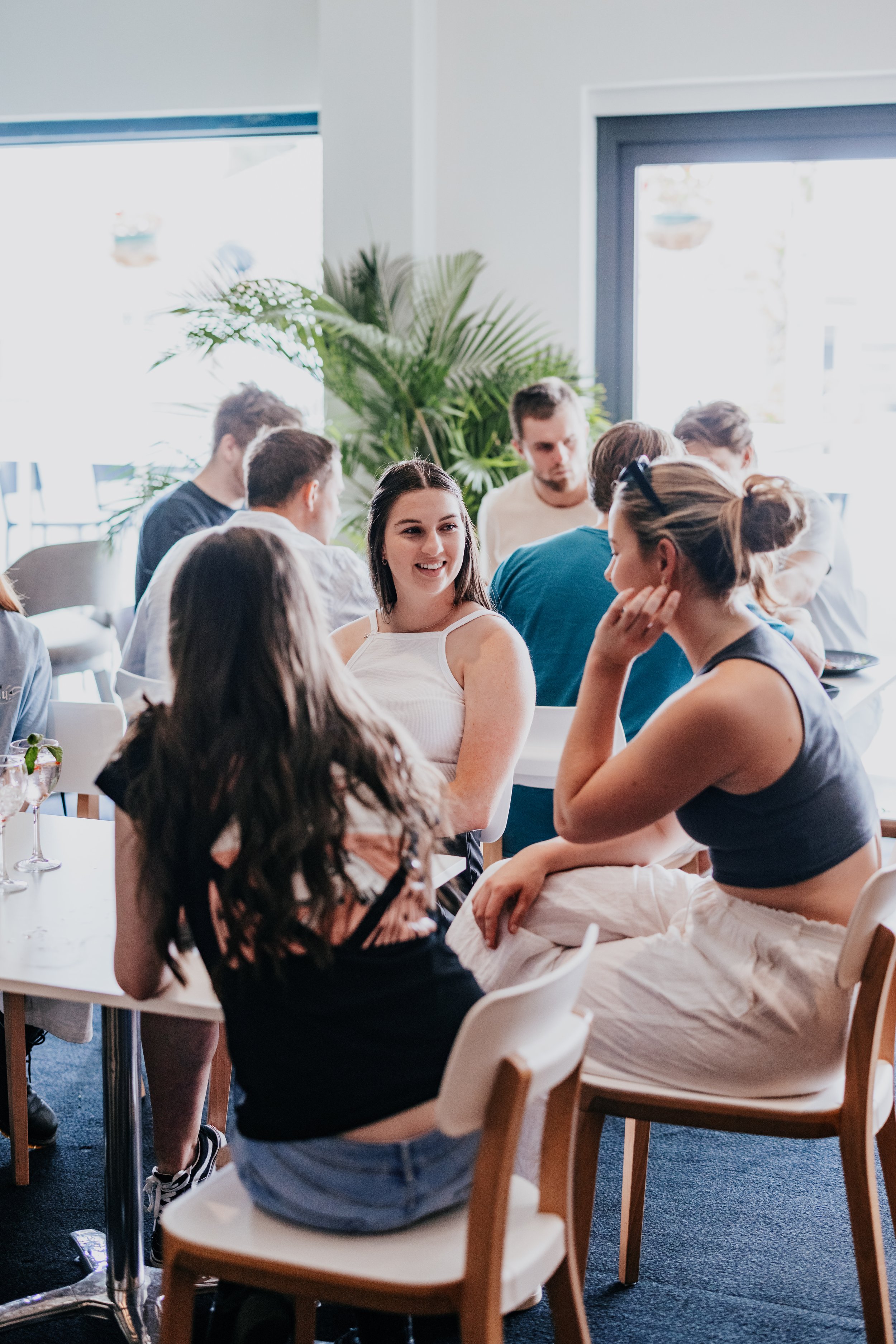 A group of young adults gathered around a table in a bright, modern cafe with large windows and indoor plants, engaging in conversation and smiling.