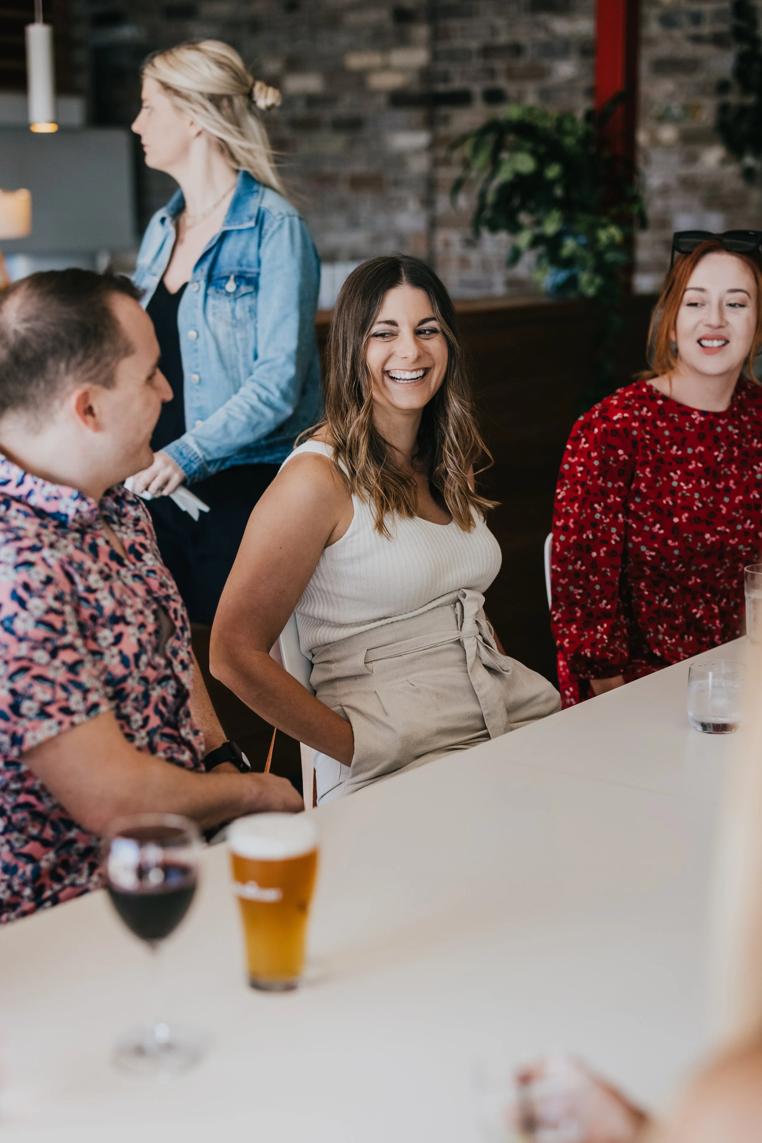 Group of friends enjoying drinks and conversation at a social gathering, with one woman smiling and the others engaged around a bar or table.