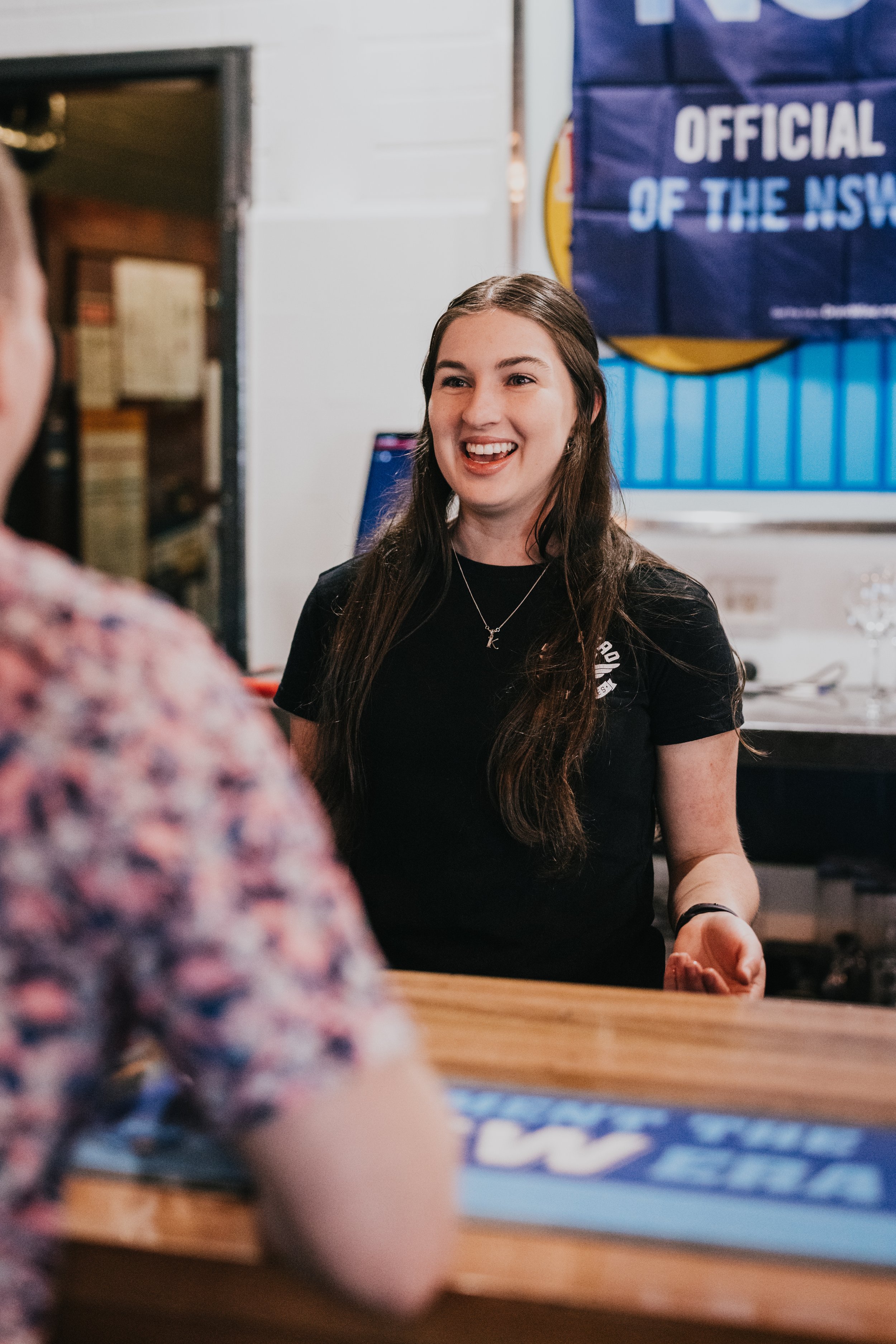 A young woman with long brown hair smiling and talking to a person in a floral shirt at a counter. A blue banner with 