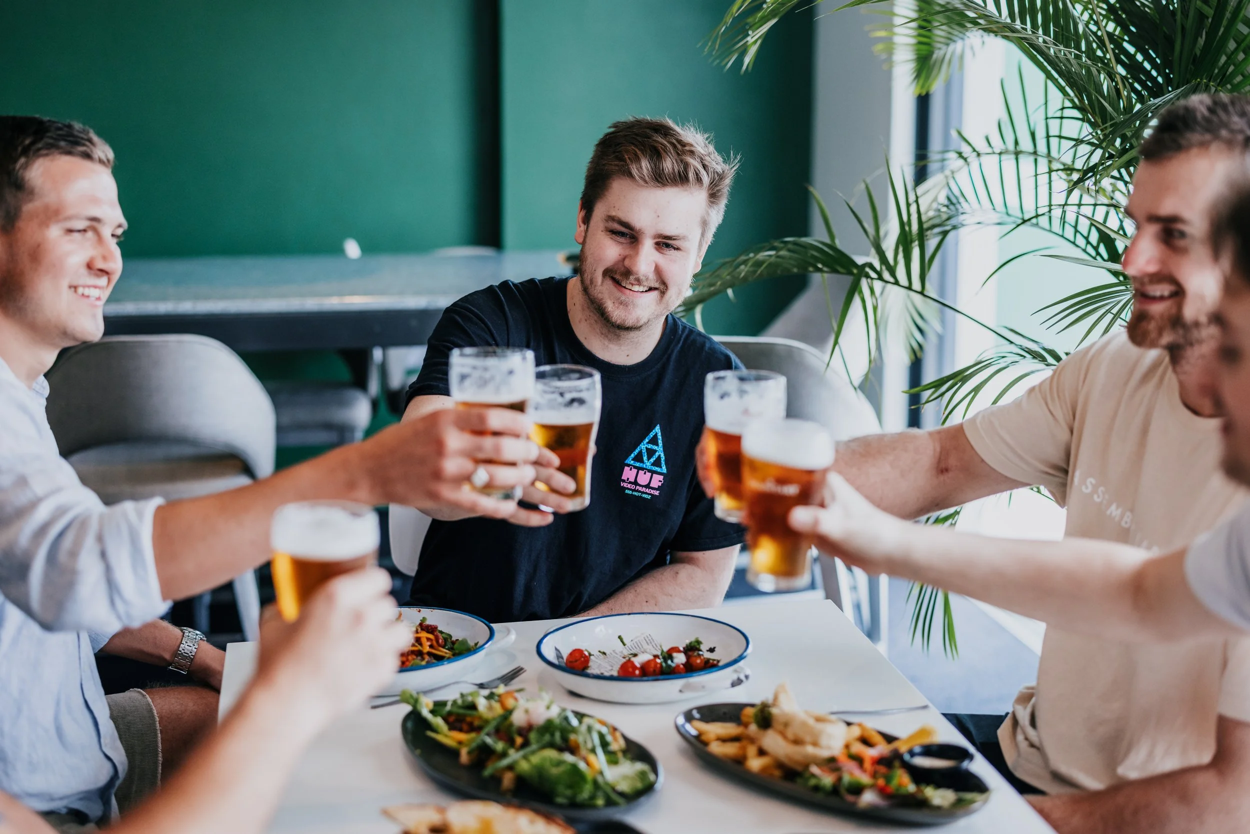 Four young men sharing drinks and toasting with glasses of beer at a dining table with salads and food in a bright indoor setting.