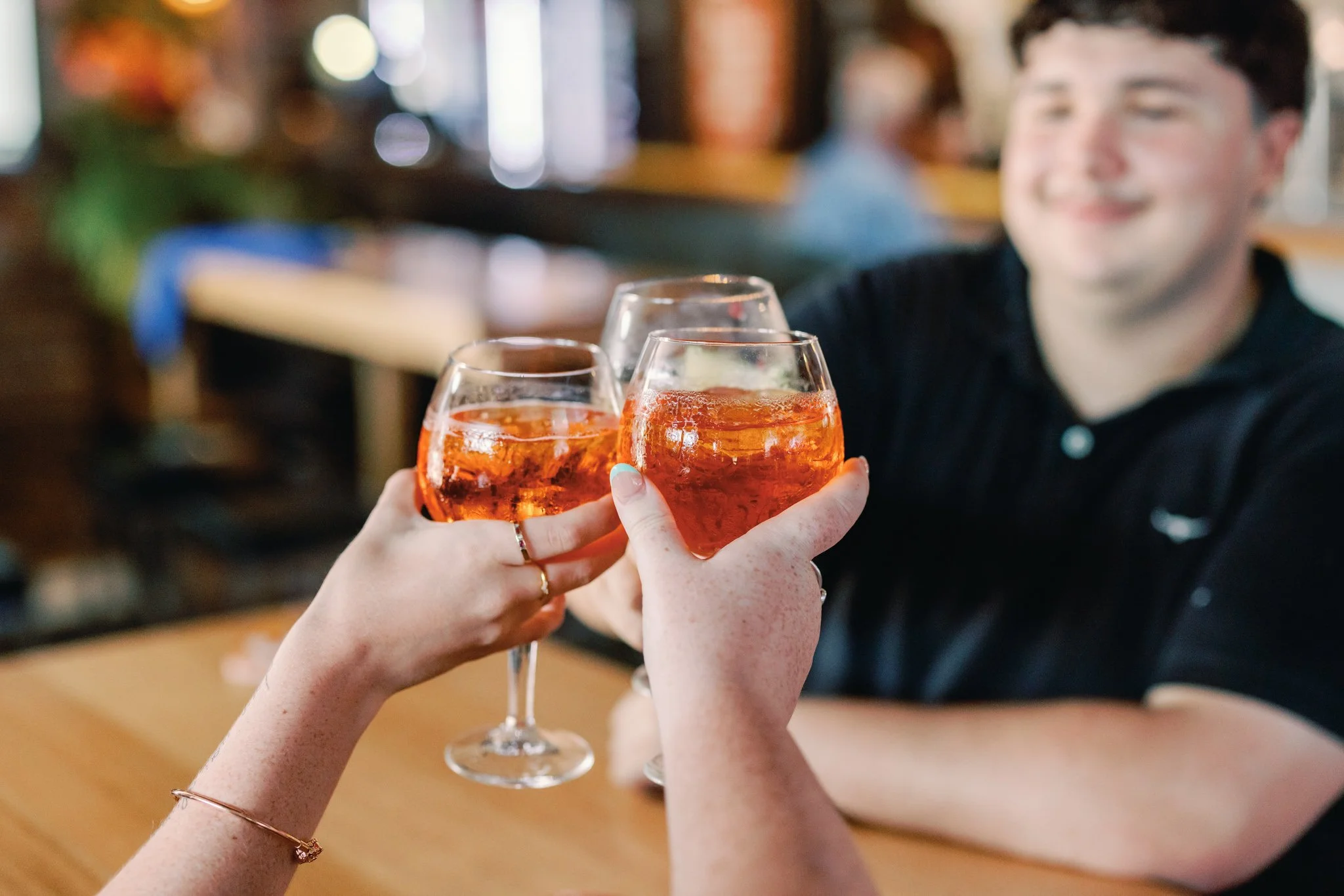 Three people clinking glasses of rosé wine in a toast at a bar or restaurant, with smiling faces and a blurred background.