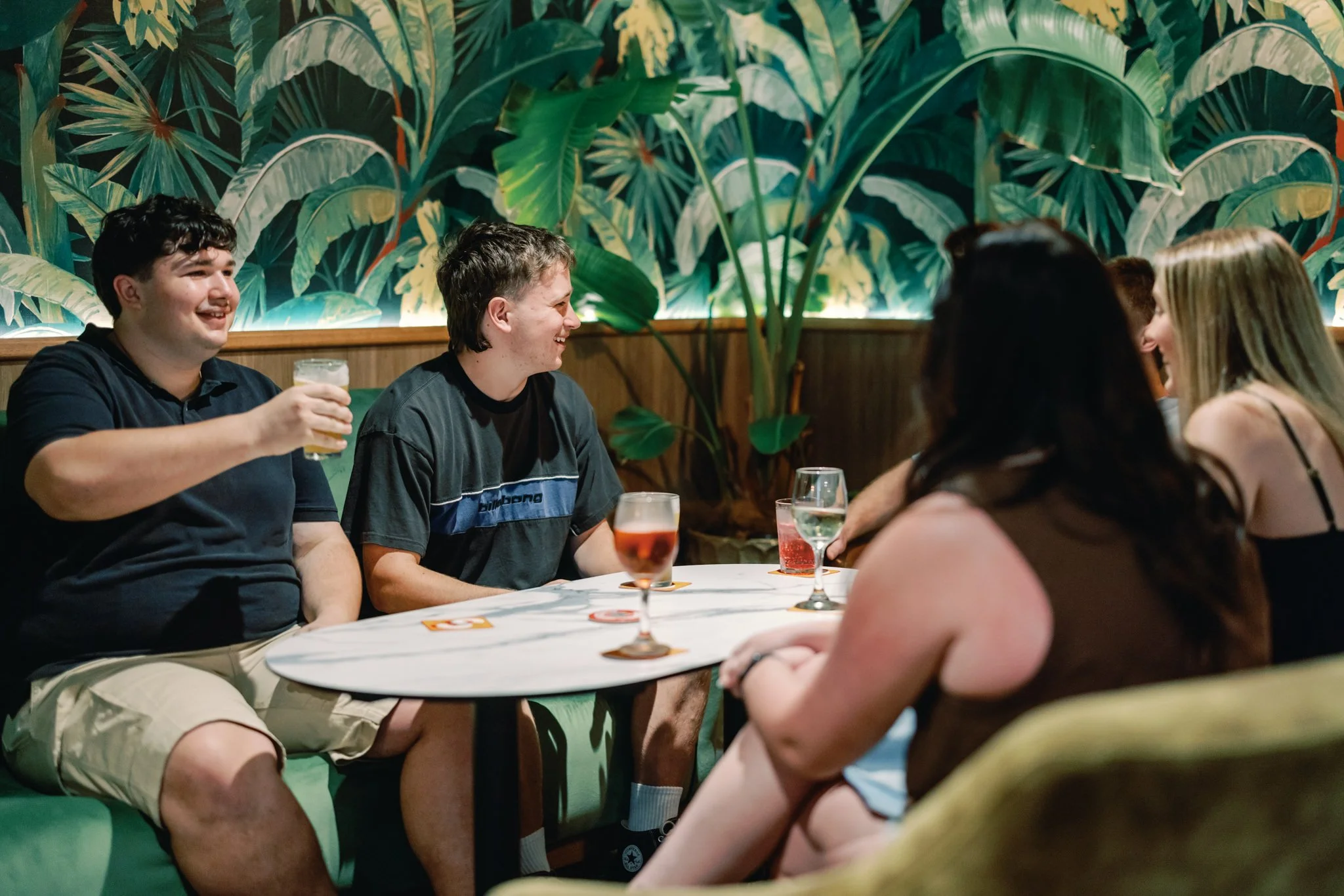 Group of young friends sitting around a table in a restaurant or bar, smiling and socializing, with drinks on the table and a tropical-themed wall in the background.