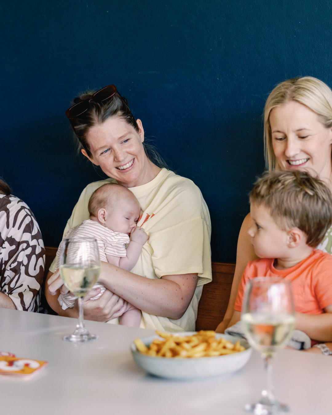 Woman holding a sleeping baby at a family gathering, people smiling, with glasses of white wine and a bowl of fries on the table.