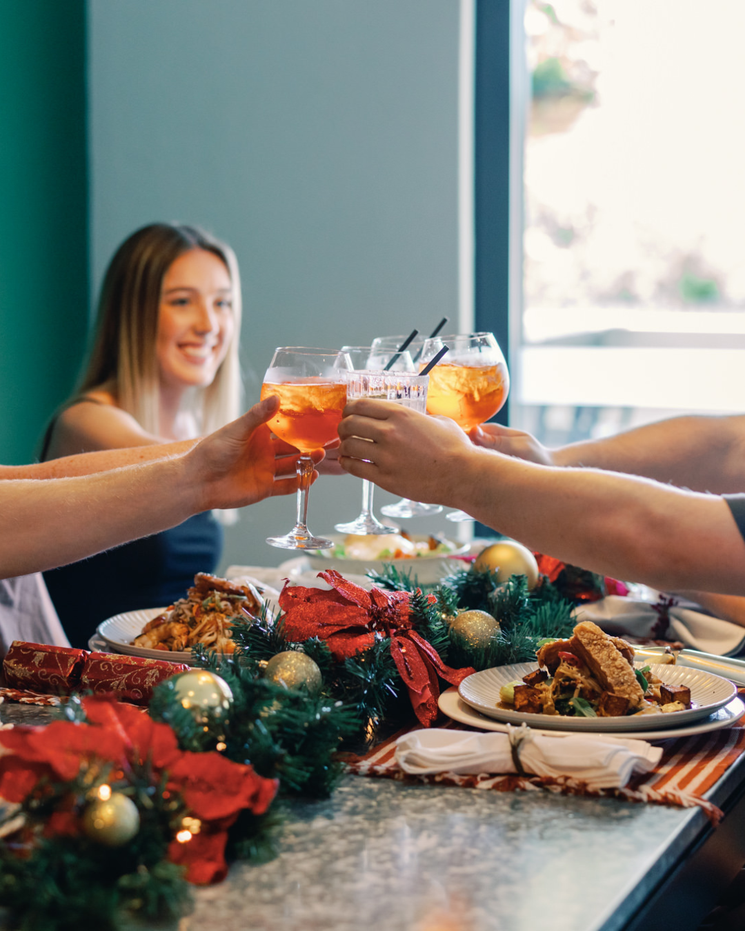 People clinking glasses of orange-colored drinks at a festive holiday dinner table decorated with poinsettia and gold ornaments.