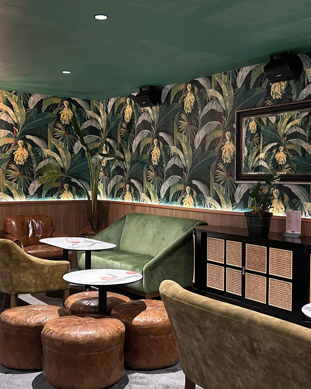 Cozy lounge area with upholstered chairs and ottomans, tropical leafy wallpaper, a potted plant on a sideboard, and framed artwork on the wall.