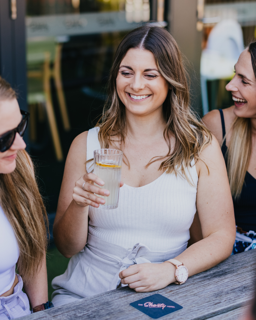 Woman holding a glass of lemonade and smiling, sitting with friends at an outdoor table.