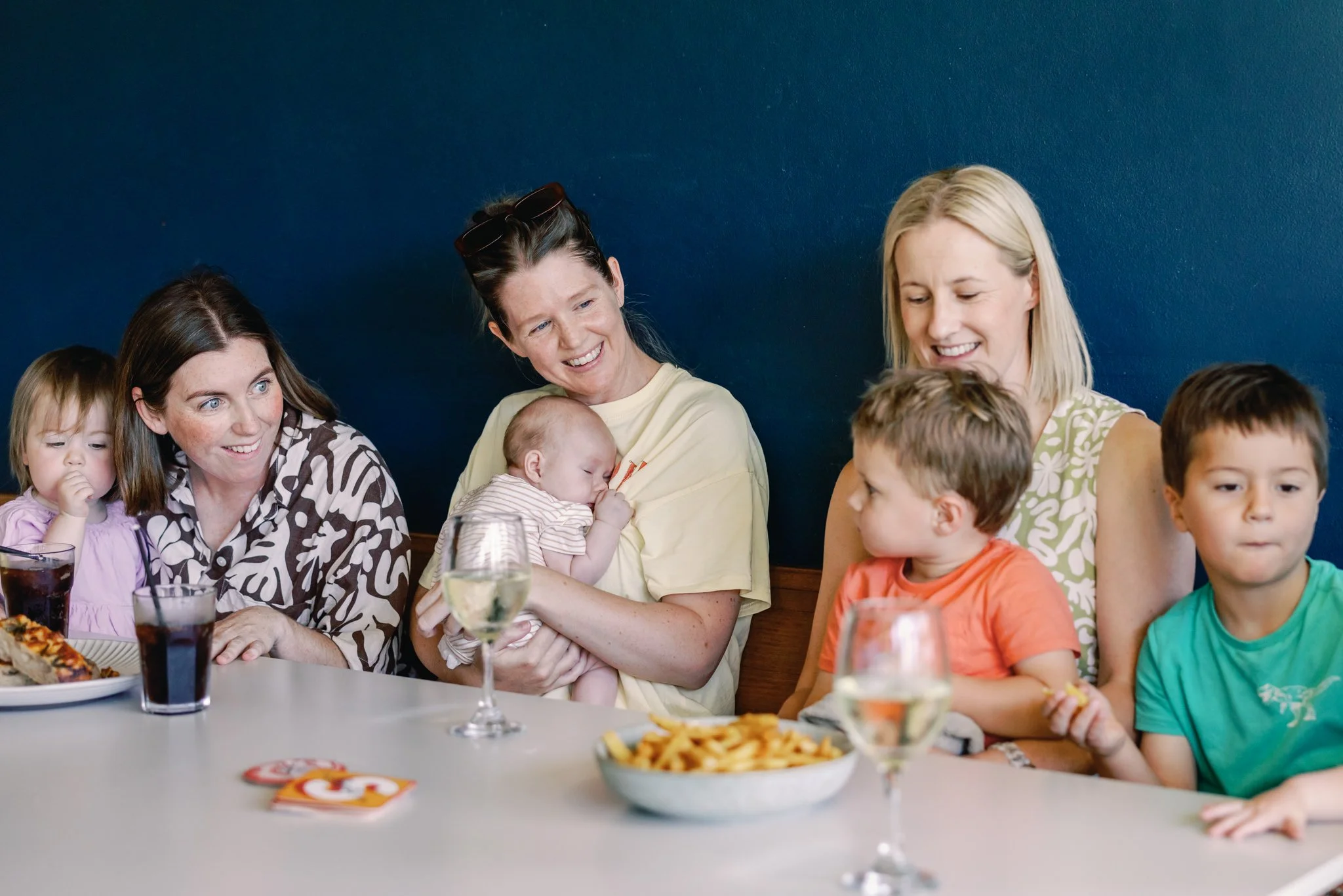 A group of women and children sitting at a dining table, enjoying food and drinks, with a dark blue wall in the background.