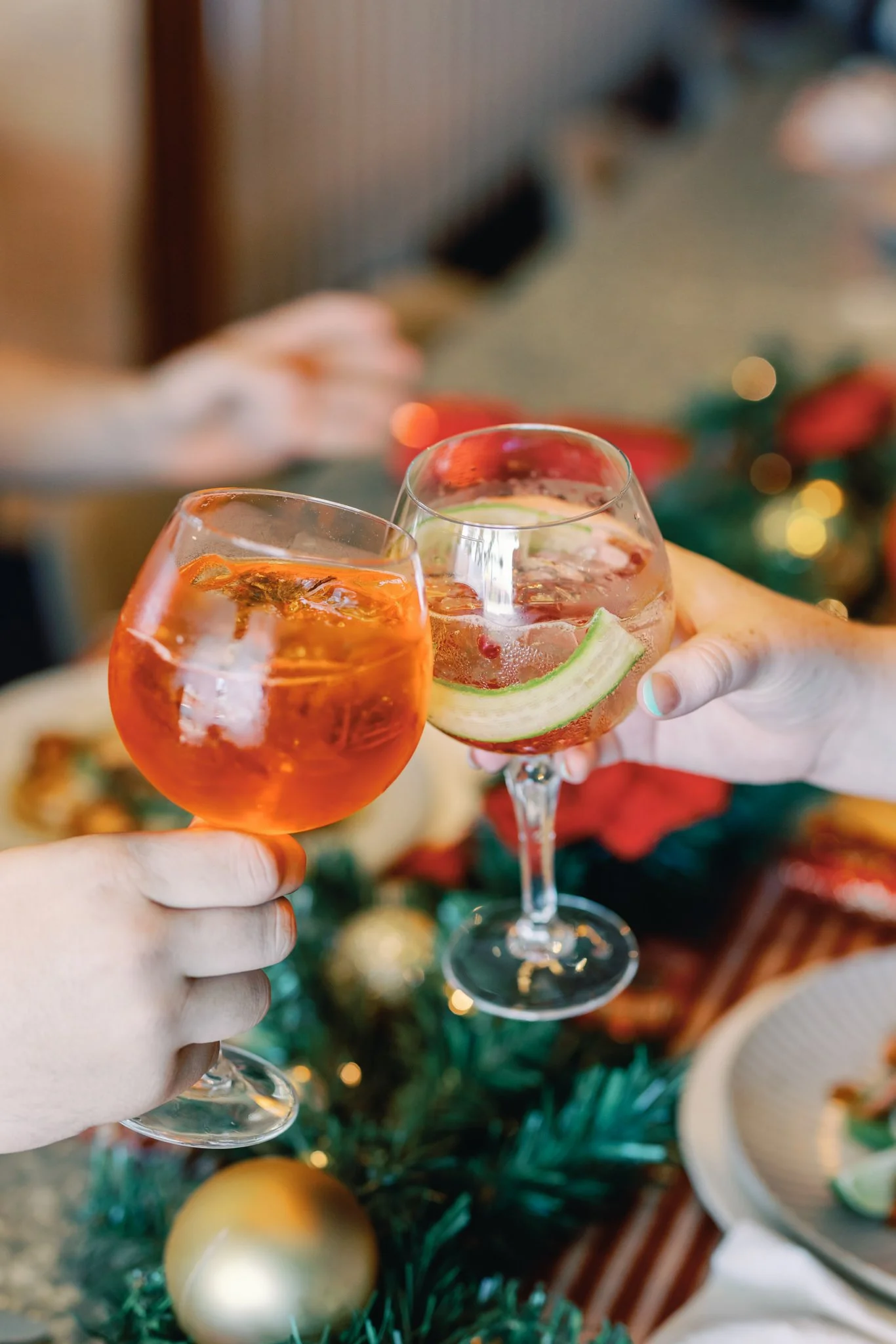 Two people toasting with glasses of festive drinks, one with a slice of lime and the other with an orange slice, holiday decorations in the background.