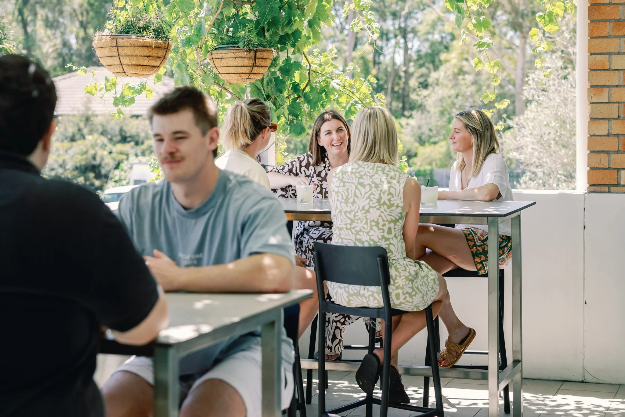 Group of friends sitting at tables on a patio, enjoying drinks and conversation in a sunny outdoor setting with hanging planters and greenery.