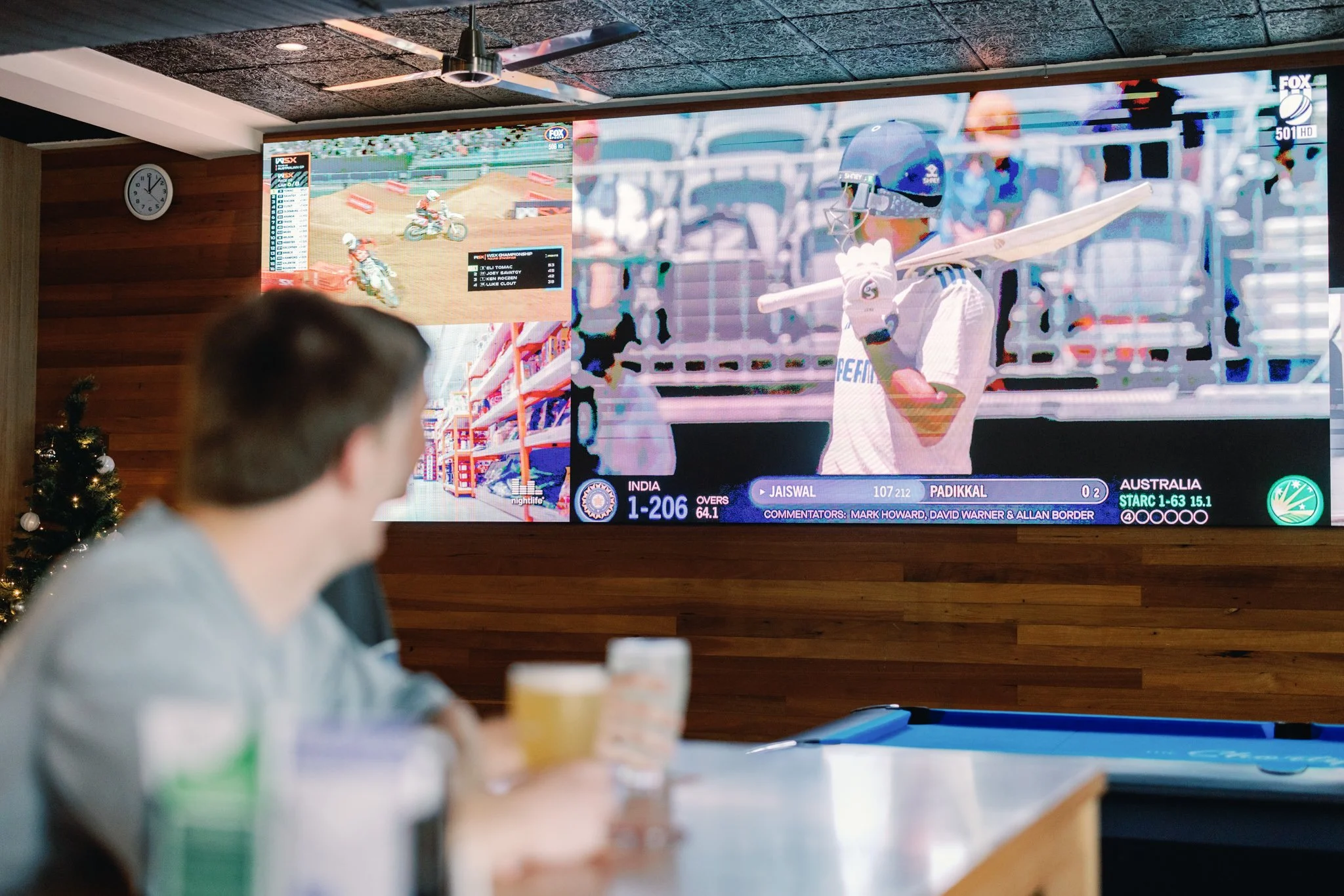 Person watching a cricket match on a large TV screen in a room decorated with a Christmas tree.