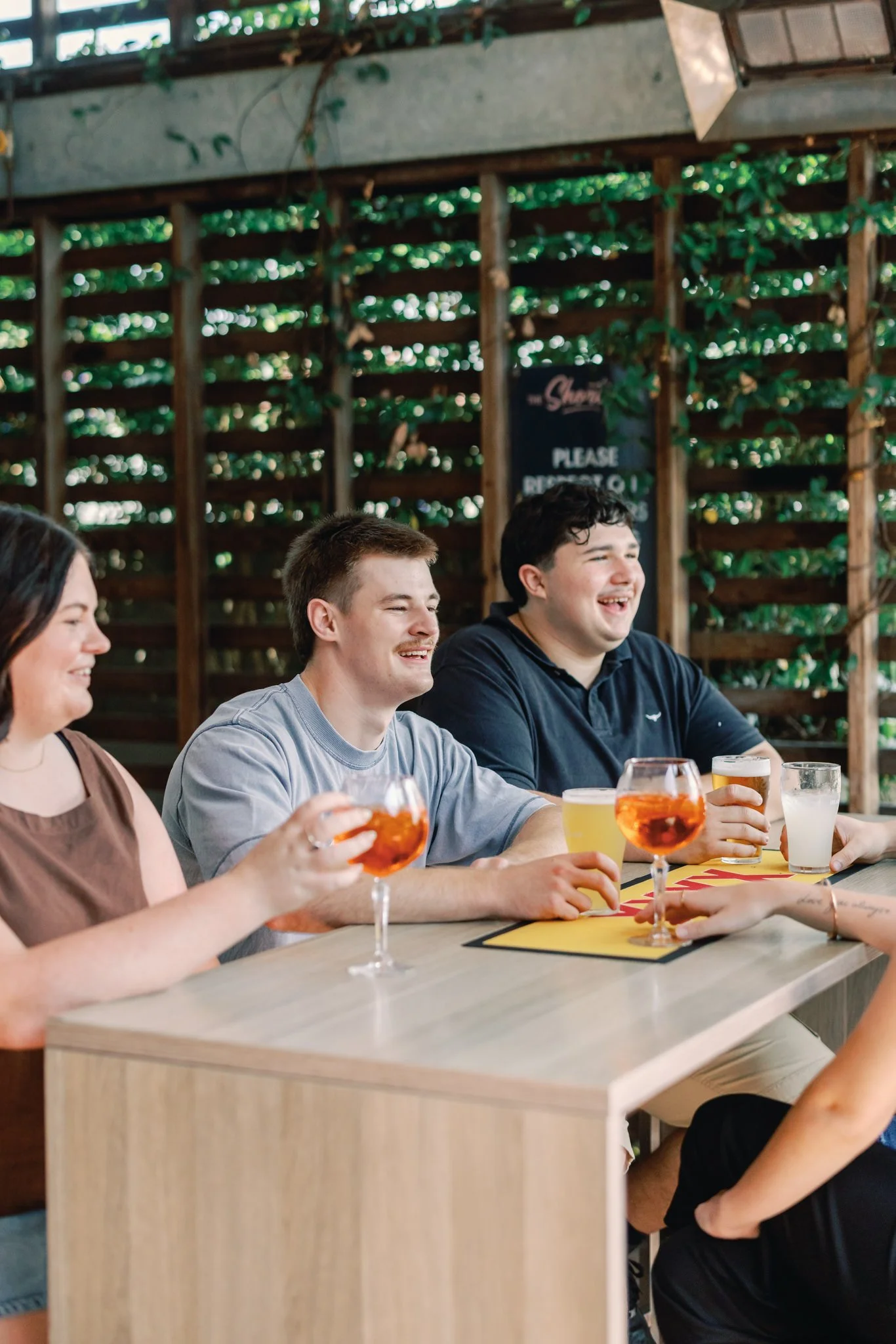 Four friends sitting at a bar table laughing and enjoying drinks, with a wooden lattice wall with greenery behind them.