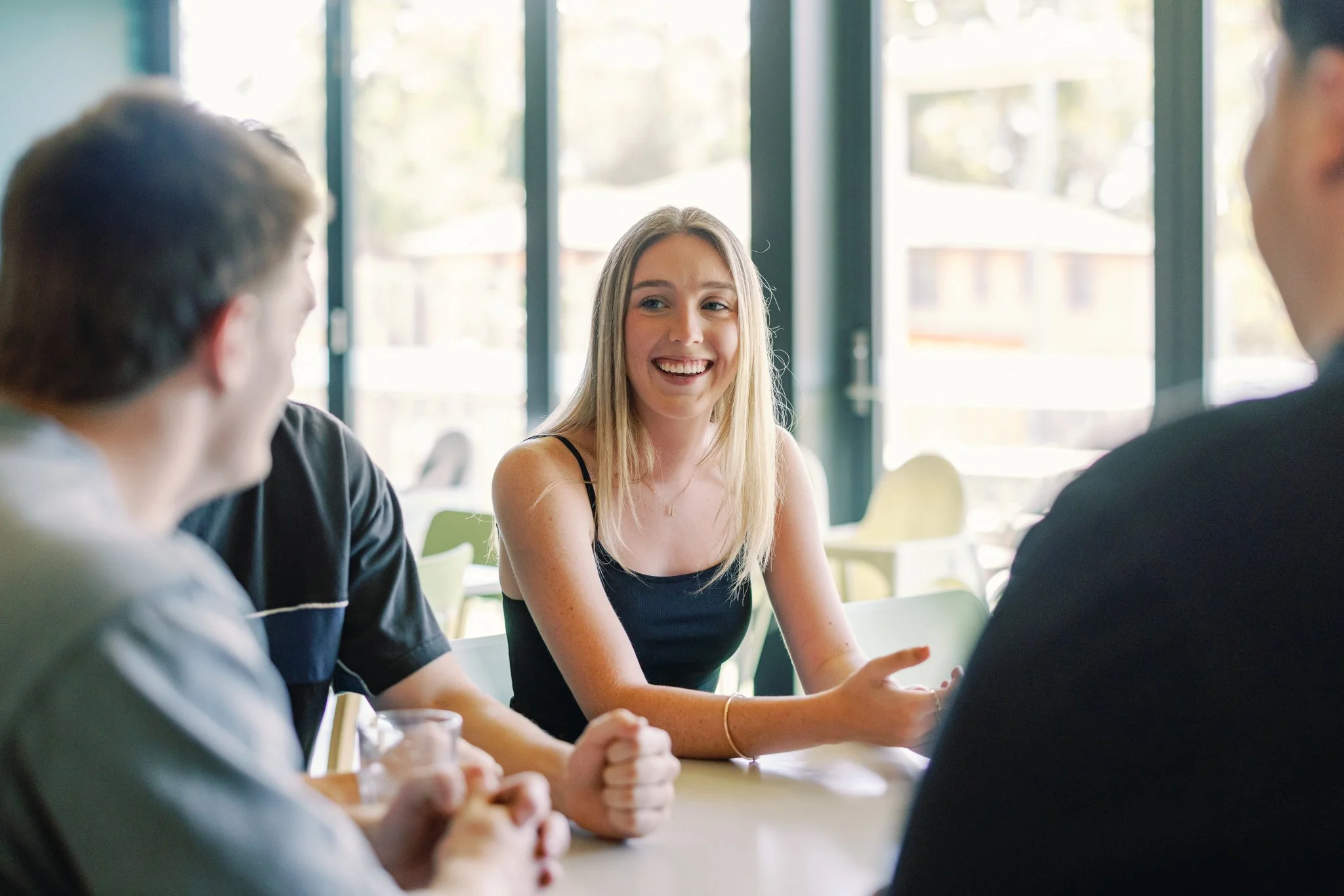 A young woman with blonde hair smiling and talking with three people at a table in a brightly lit cafe or restaurant.