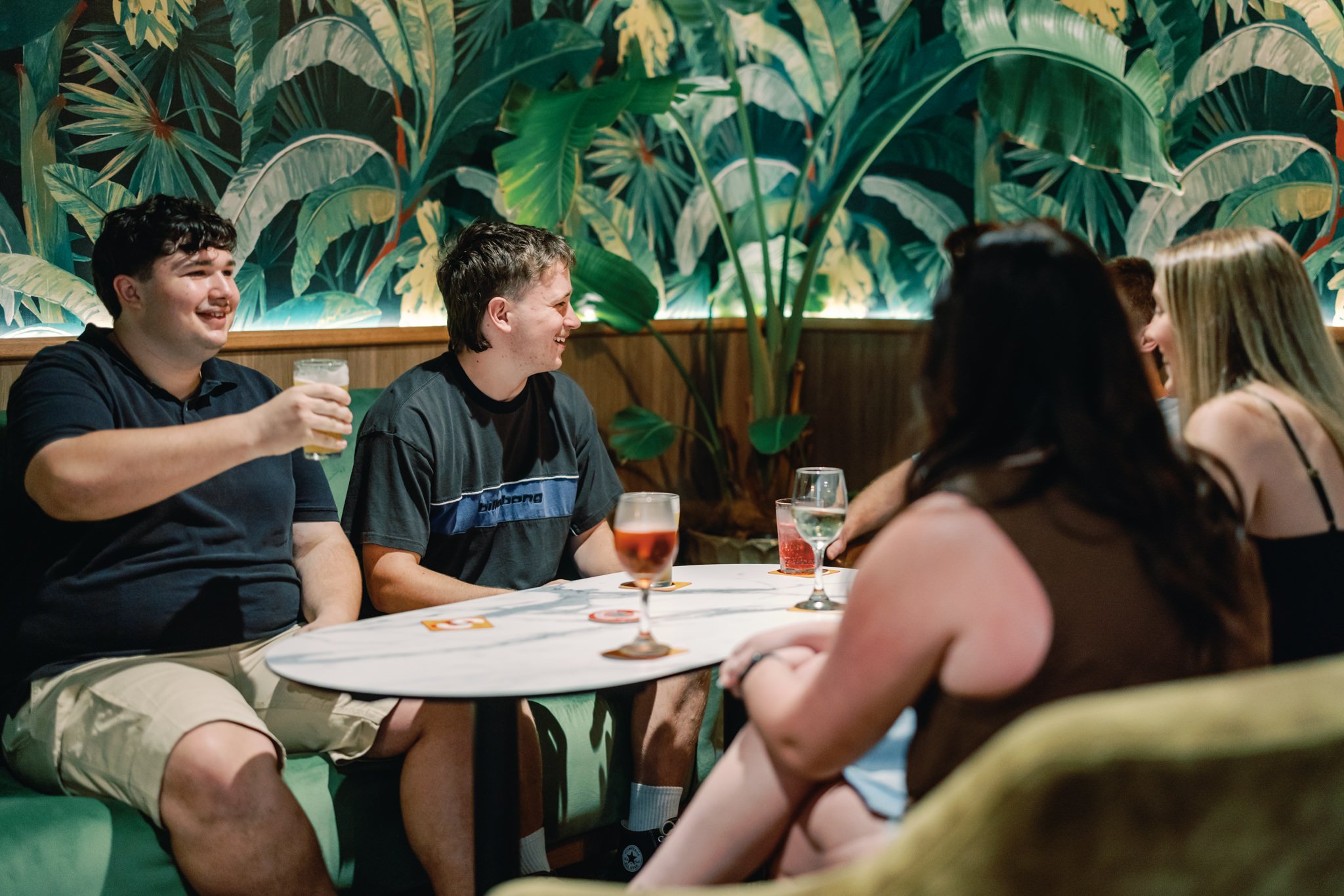 Group of young people sitting around a table in a restaurant, chatting and laughing, with drinks on the table and tropical plant wallpaper in the background.
