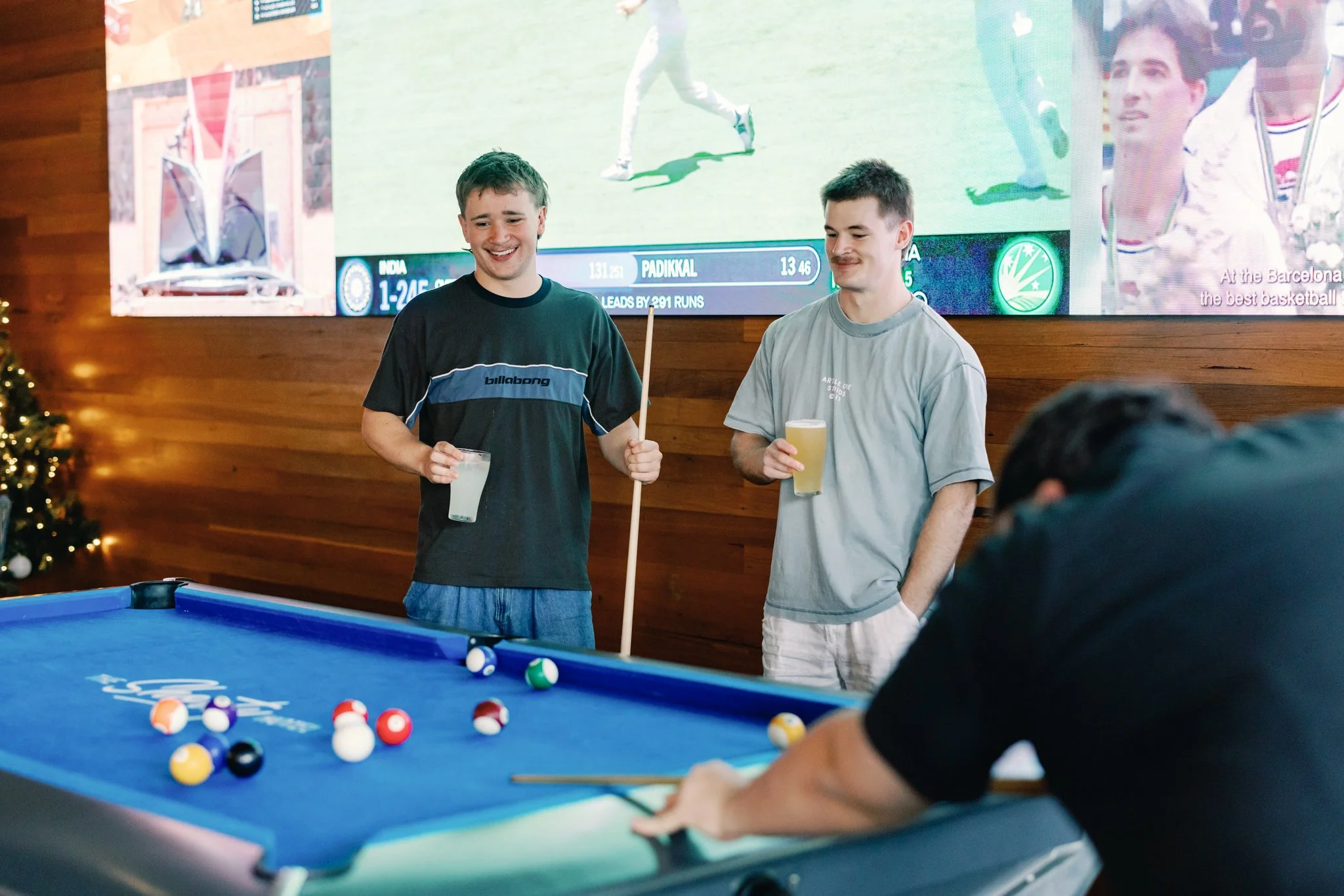 Two young men standing by a pool table, smiling and holding drinks, while another person is leaning over the table aiming to take a shot. In the background, a large screen displays a cricket match with scores and player information.