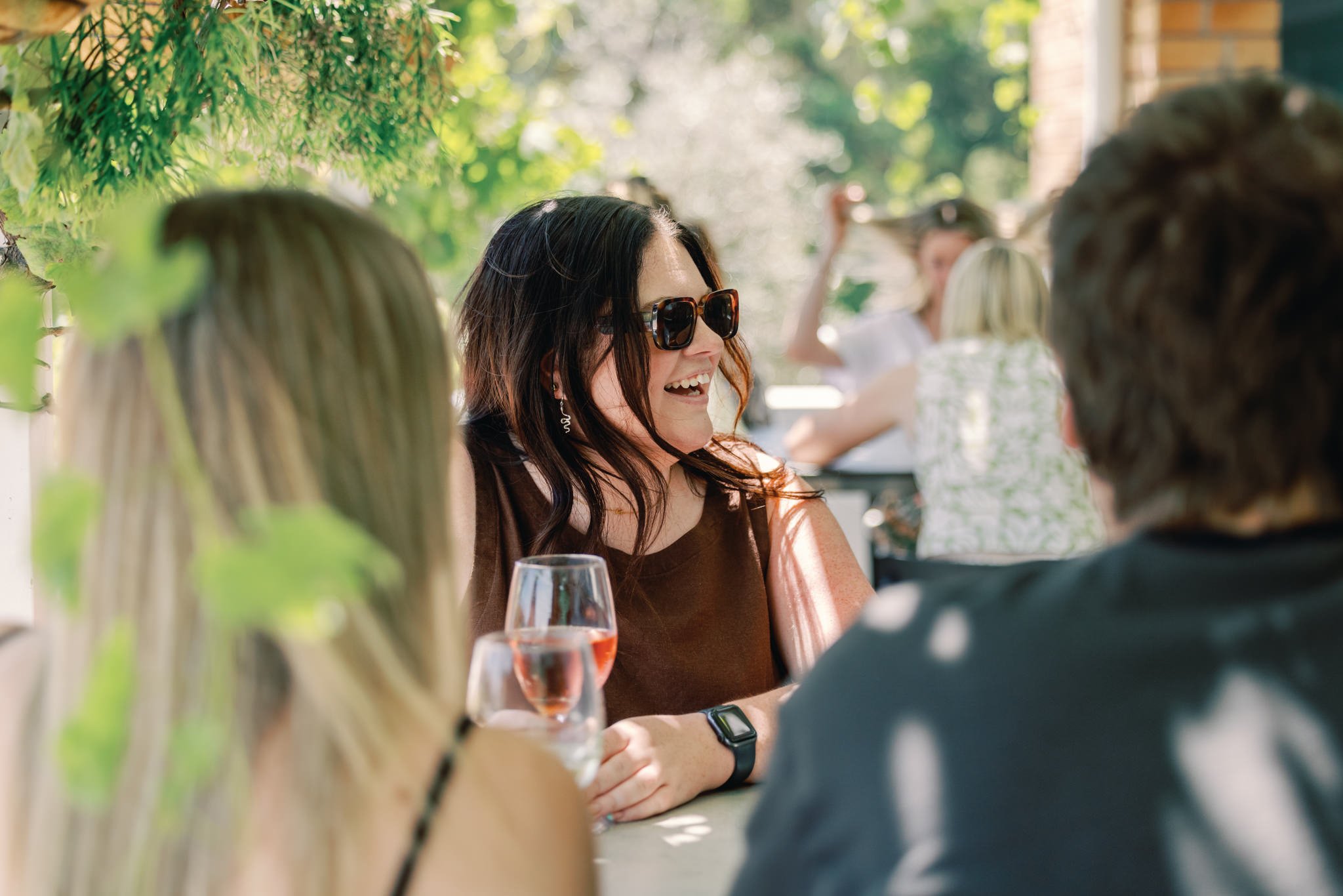 Group of people socializing outdoors at a table on a sunny day, with greenery and a brick building in the background.