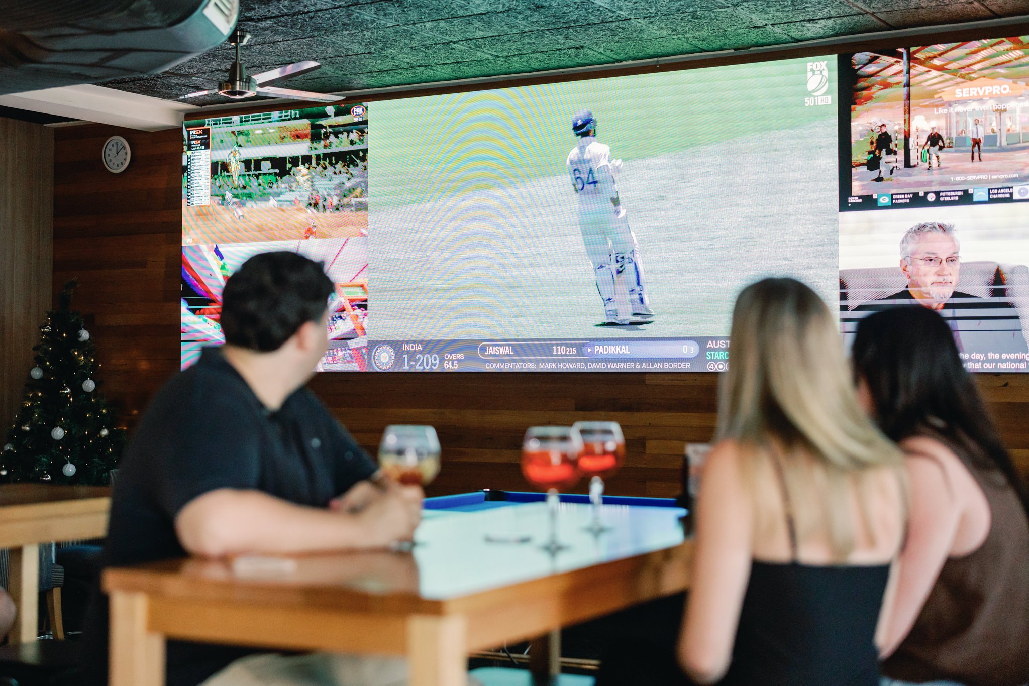 People watching a cricket match on a large screen in a bar with drinks on the table. The screen shows a player standing on the field and other broadcasts.