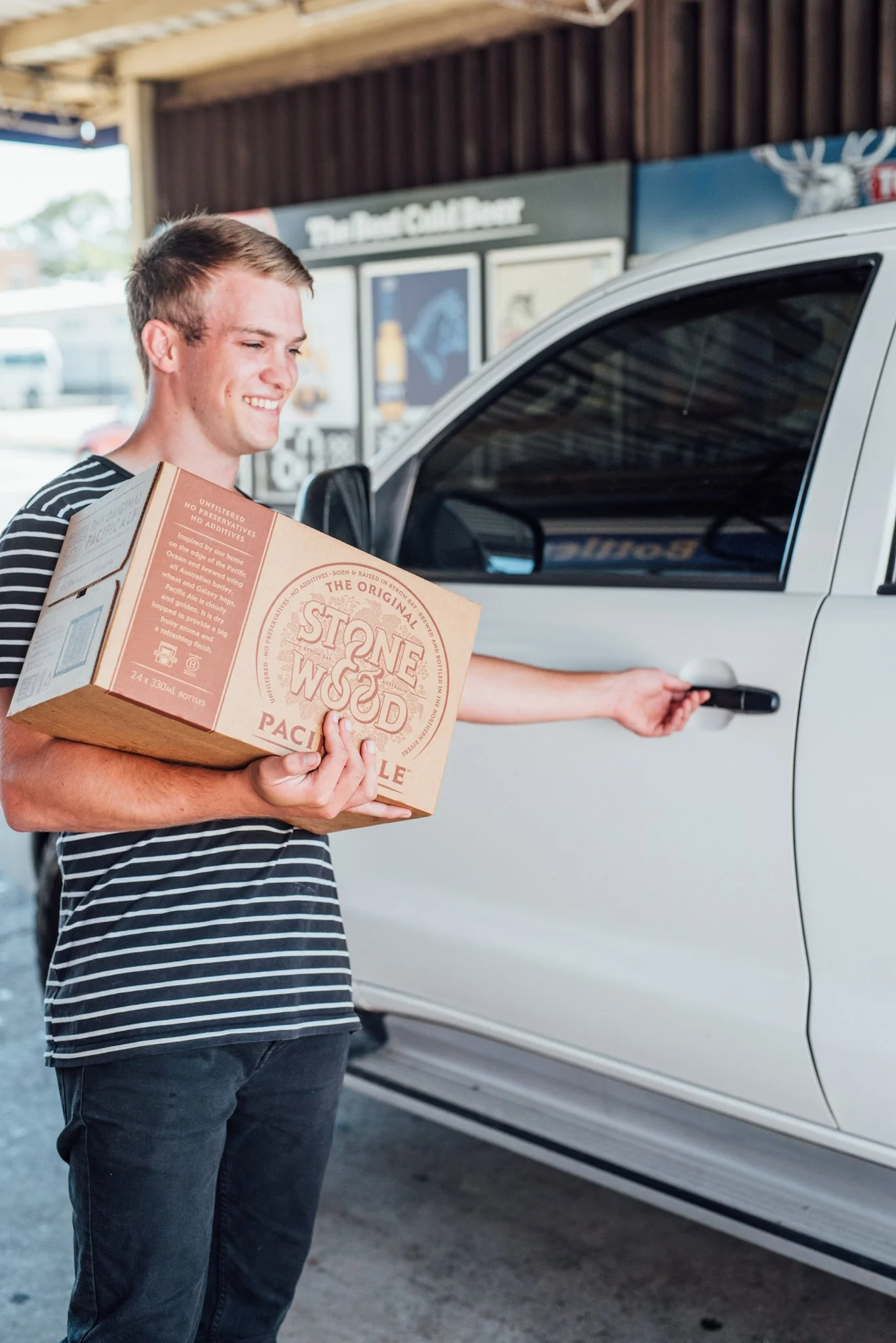 A smiling young man holding a box of Stone & Wood Pacific Ale beer bottles while opening the car door of a silver vehicle.