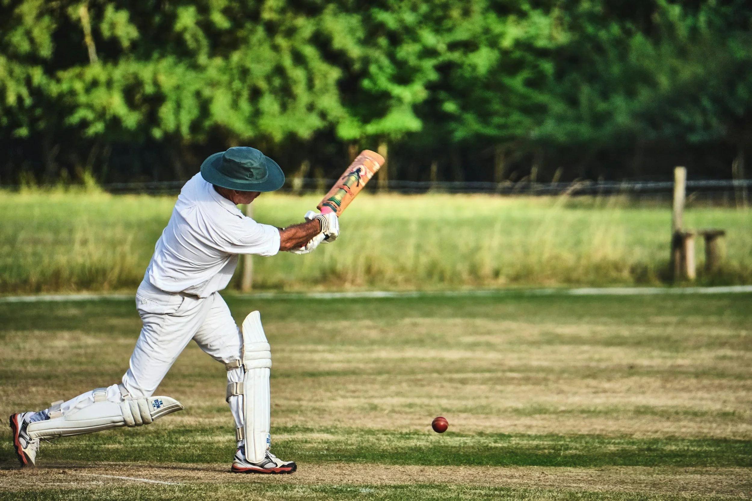 A man in white cricket gear and a large hat is batting on a cricket field, preparing to hit a red cricket ball.
