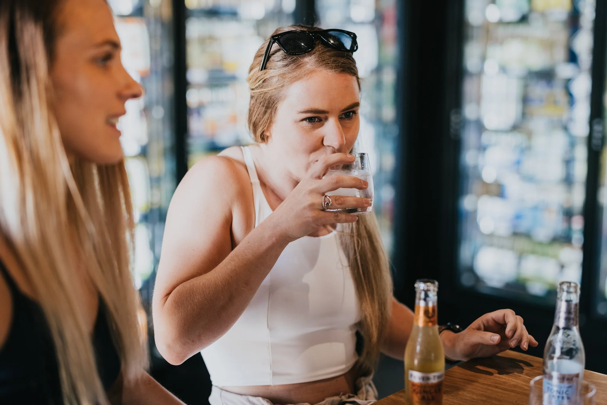 A young woman with sunglasses on her head is drinking a glass of water at a table in a cafe, with another woman with long blonde hair near her, and bottles of beer or soda on the table.