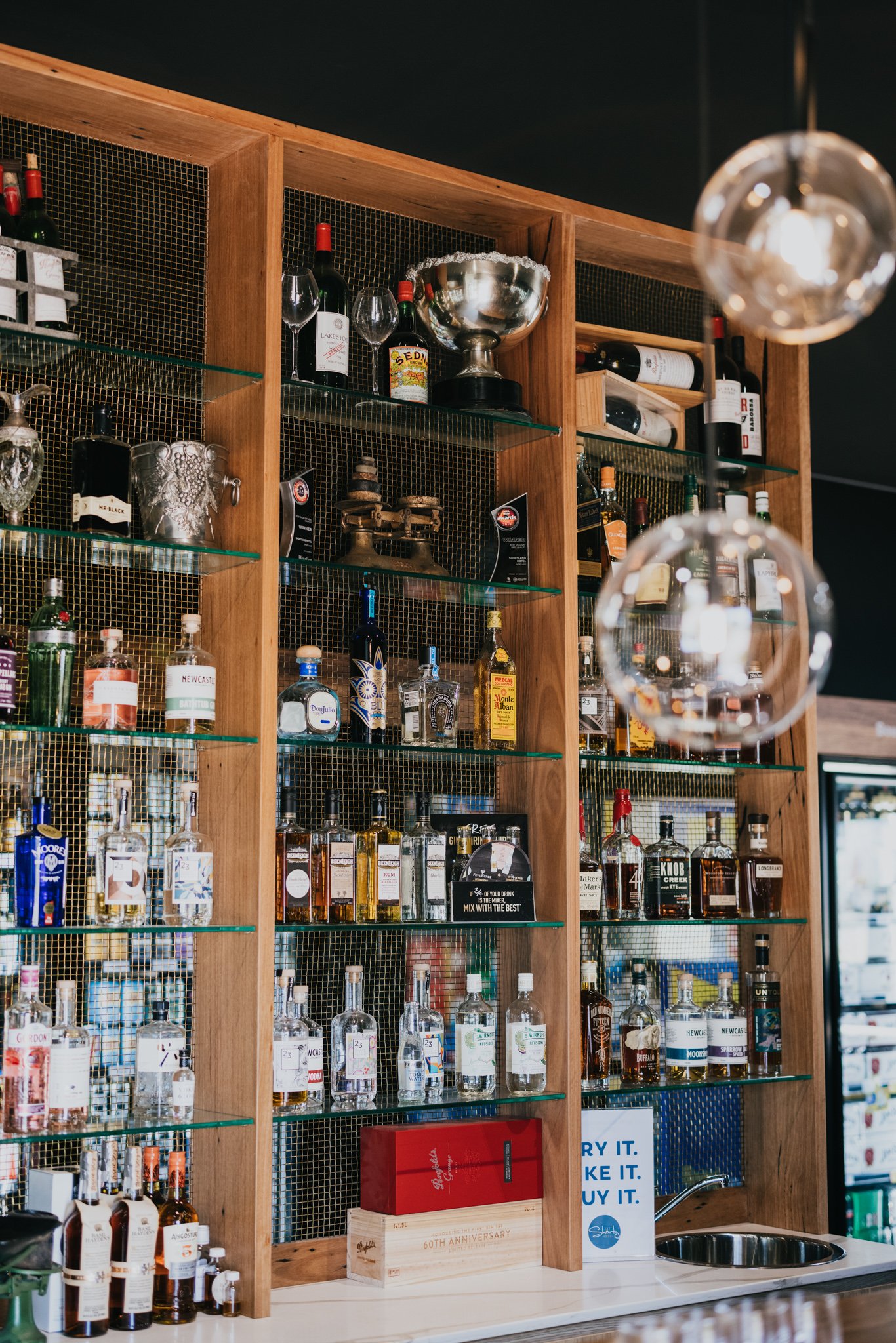 A bar with wooden shelves holding various bottles of liquor, glasses, and decorative silver items, with a countertop and a sink.