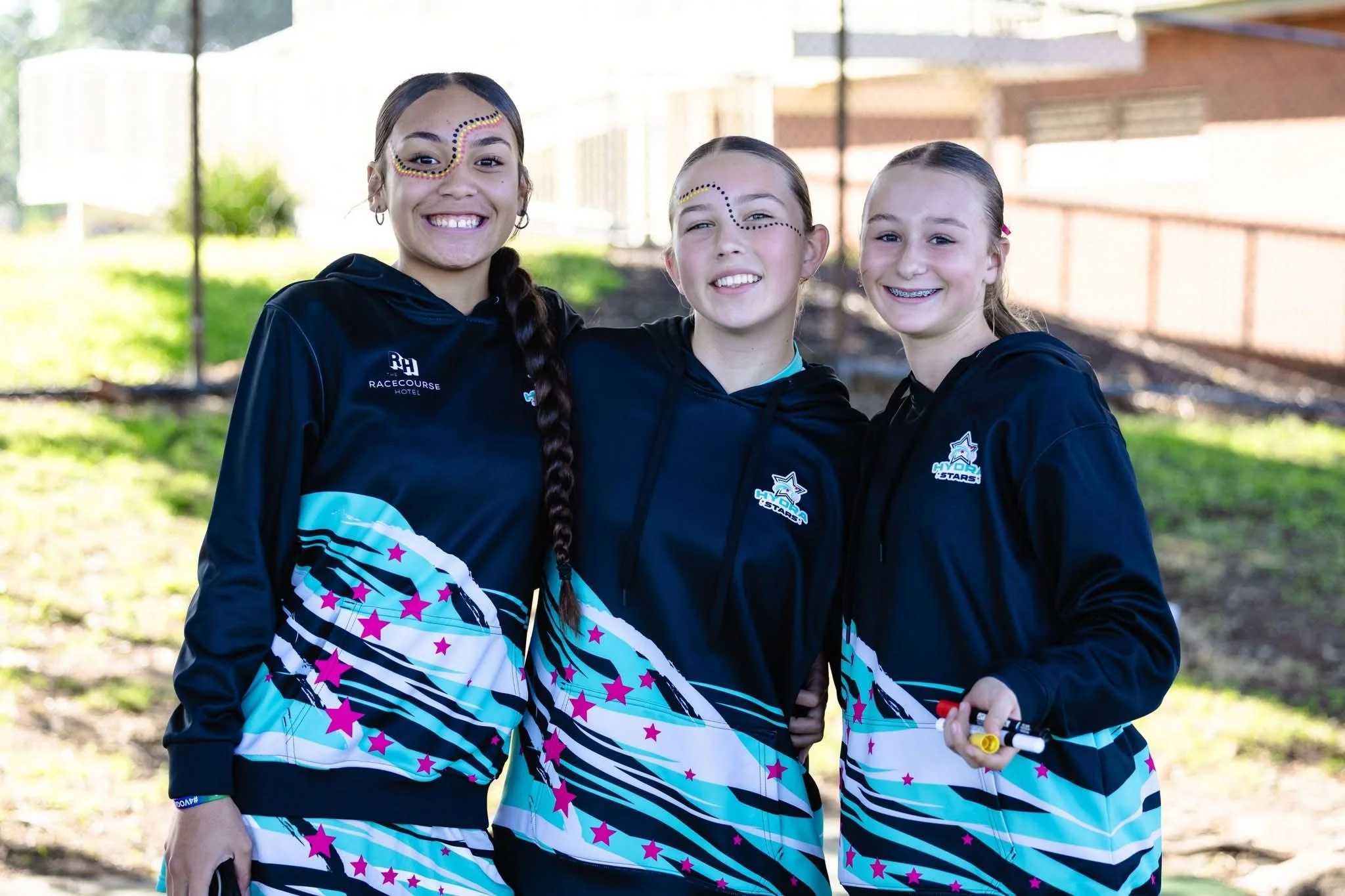 Three young girls standing outdoors, smiling, wearing black and blue sports jackets with blue and pink star patterns, and face paint designs. One girl is holding a marker.
