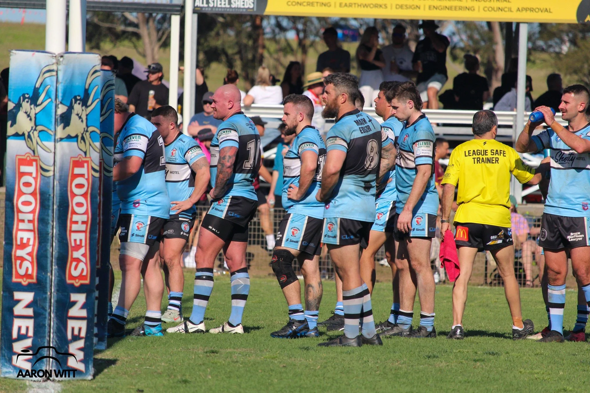 Australian rugby players in blue jerseys stand in line on the field during a game, with a referee in a yellow jersey labeled 'League Safe Trainer' nearby. Spectators sit behind a fence under a shaded area in the background.