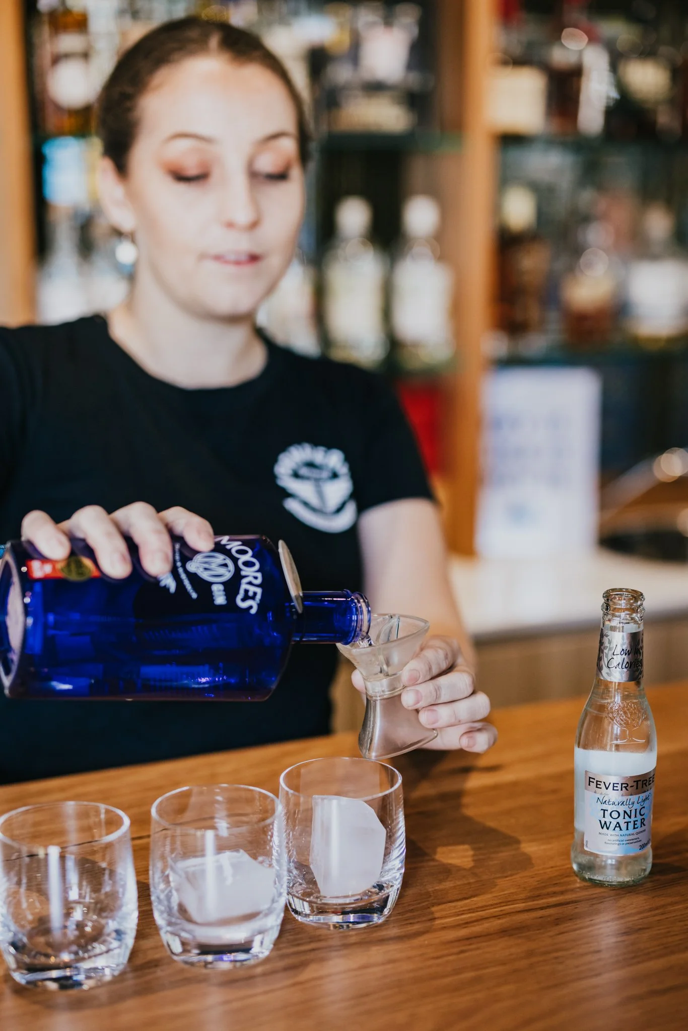A woman pouring a blue liquid from a blue bottle into a small glass with a large ice cube on a wooden bar counter. There are two other glasses with ice cubes and a bottle of tonic water labeled 'Fever-Tree' nearby.