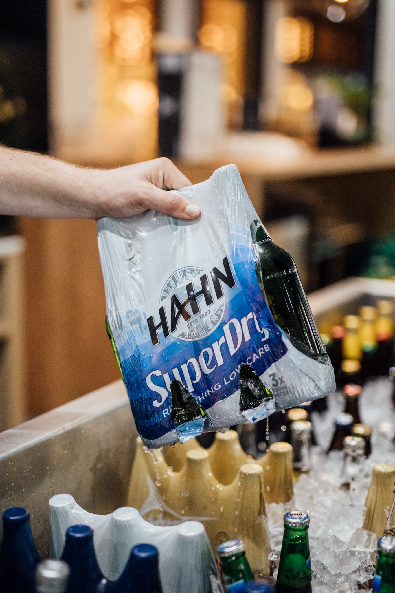 Someone's hand holding a six-pack of Haan SuperDry beverages, cooling in an ice-filled container with various bottled drinks.