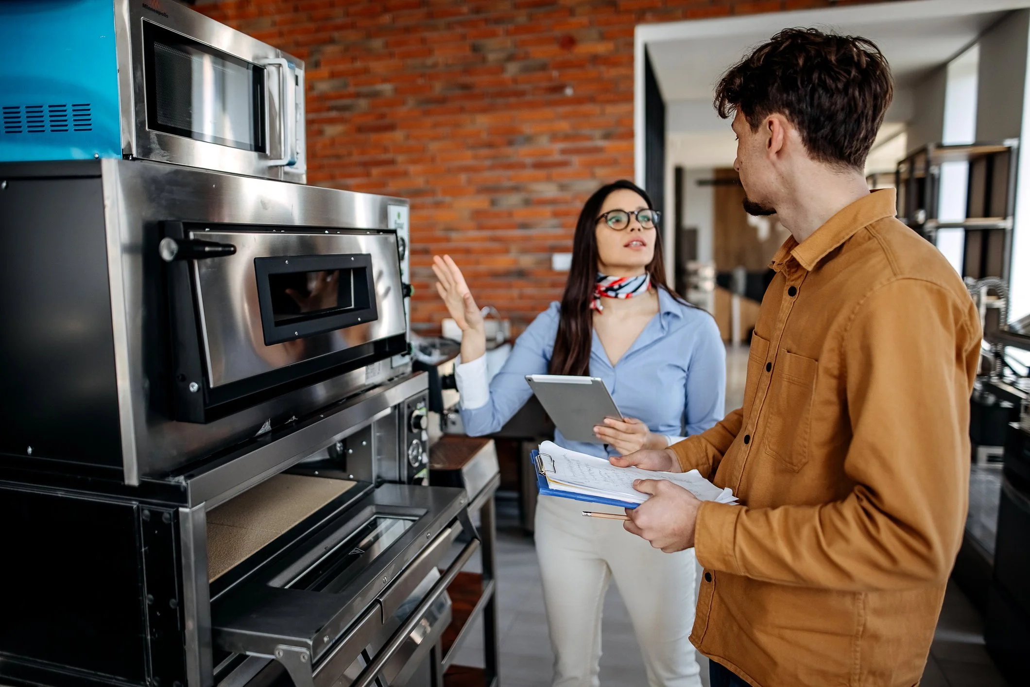 Two people having a discussion in a commercial kitchen, with one woman gesturing towards an industrial oven and holding a tablet, while the man holds a clipboard and appears to be listening.