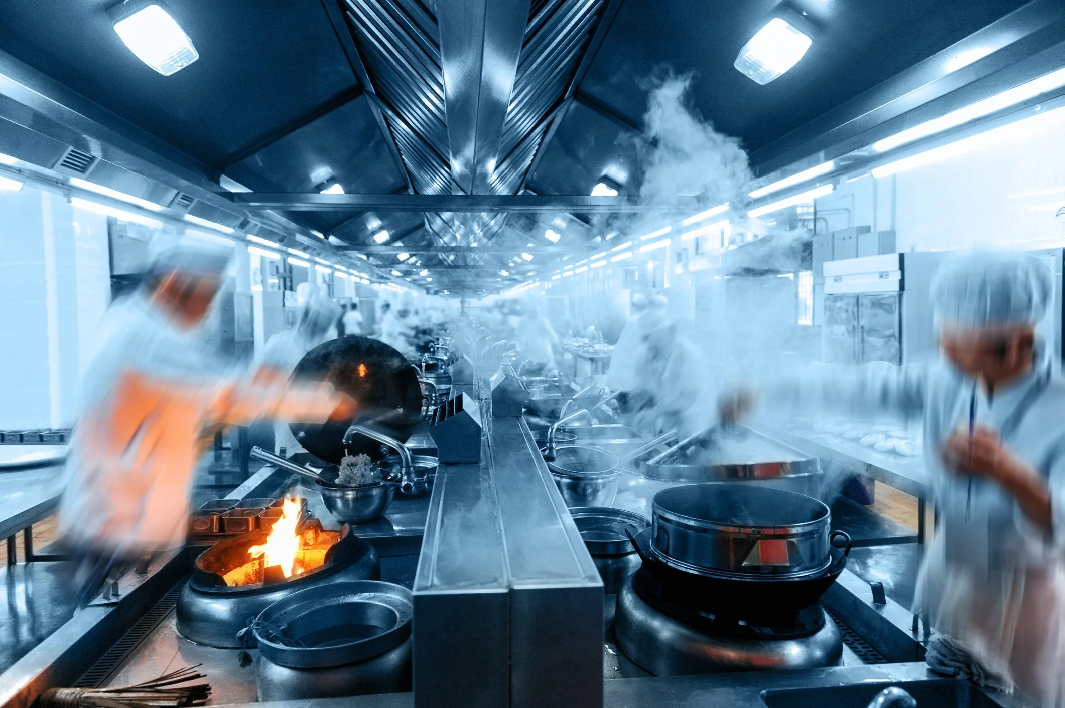 A busy commercial kitchen with chefs wearing white uniforms and hats, cooking at multiple stoves with flames and steam, in a well-lit, industrial-style space.