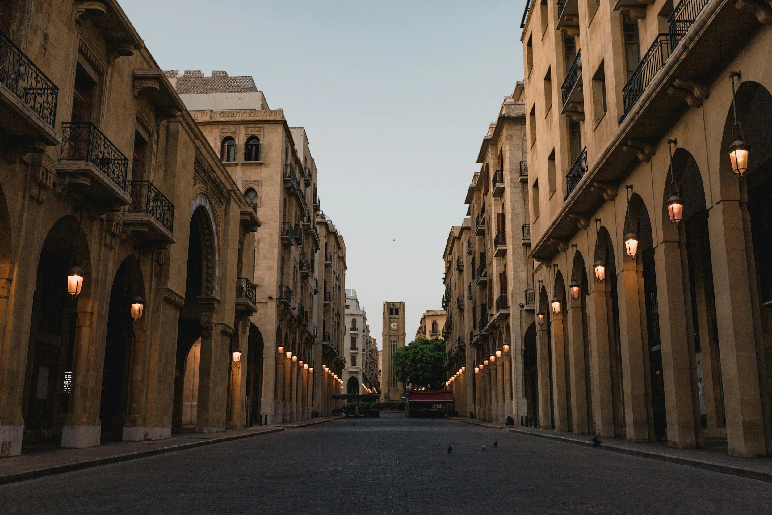 Beirut downtown with historic buildings on both sides, lit street lamps, and a clock tower in the distance under a clear sky.