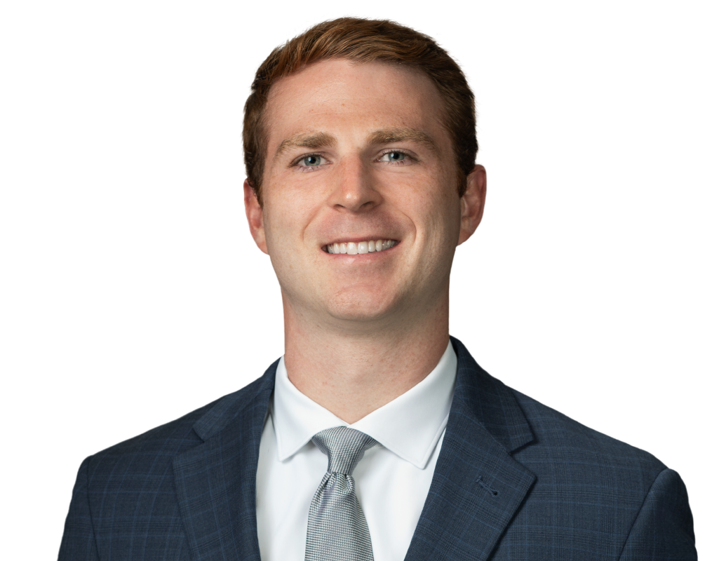 A smiling man with light brown hair, wearing a dark blue suit, white shirt, and light grey patterned tie.