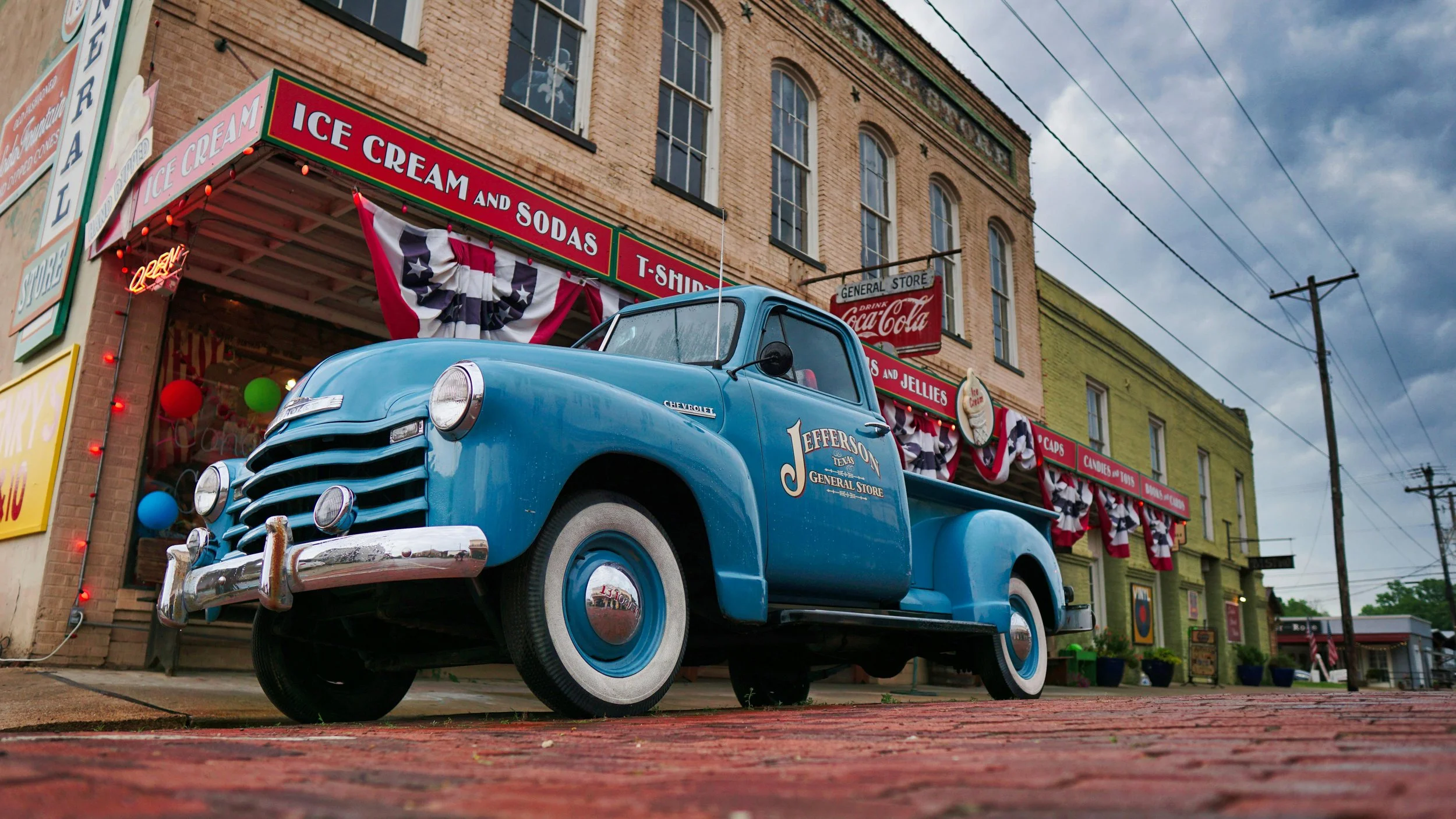 A vintage blue Chevrolet truck parked in front of a brick general store decorated with patriotic red, white, and blue streamers and balloons. The store has signs advertising ice cream, sodas, and jelly, with a Coca-Cola sign hanging outside. The sky is cloudy.