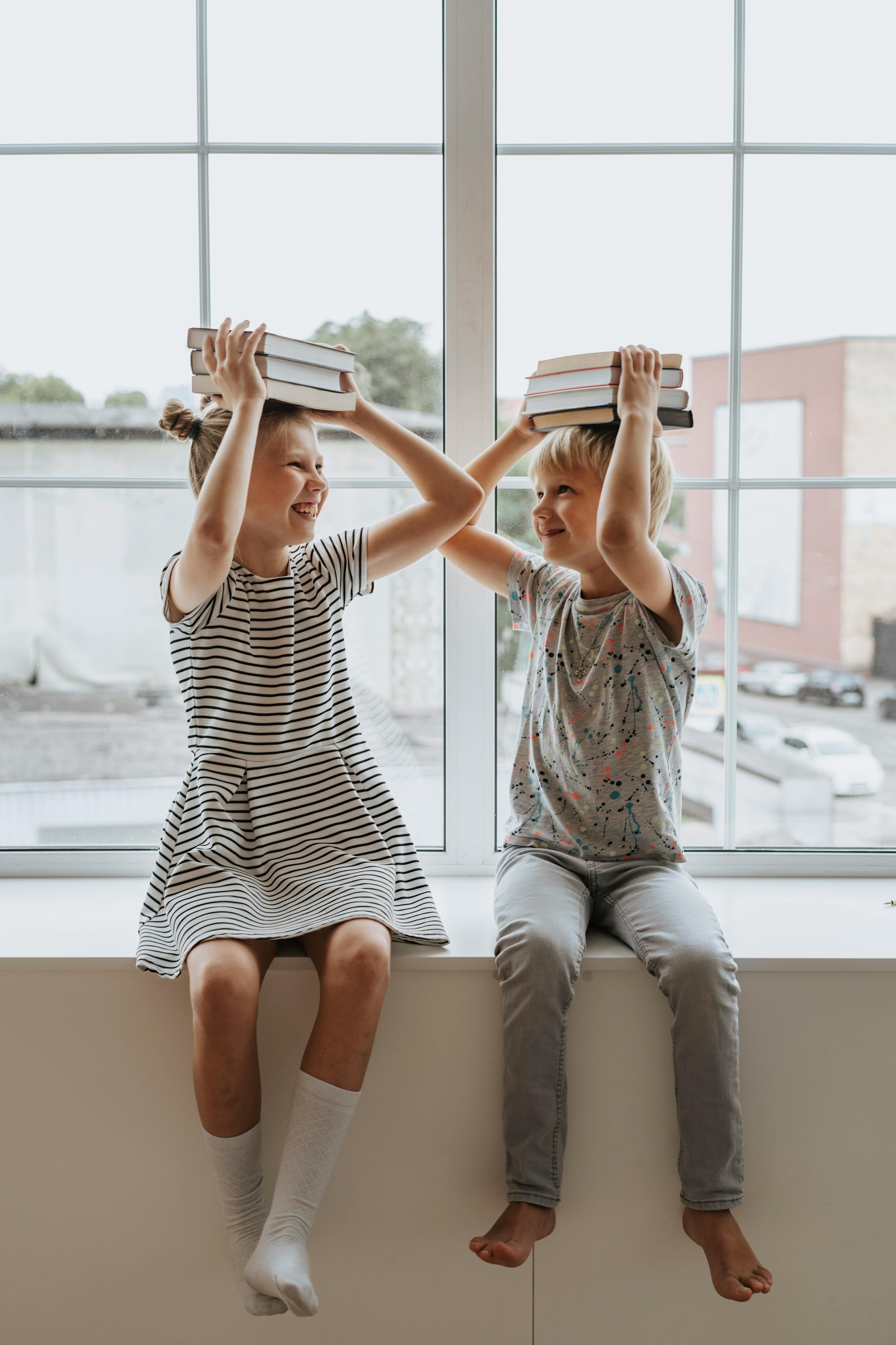 Two children, a girl and a boy, sitting on a wide window ledge, each holding a stack of books on their heads, smiling and engaging in playful interaction.