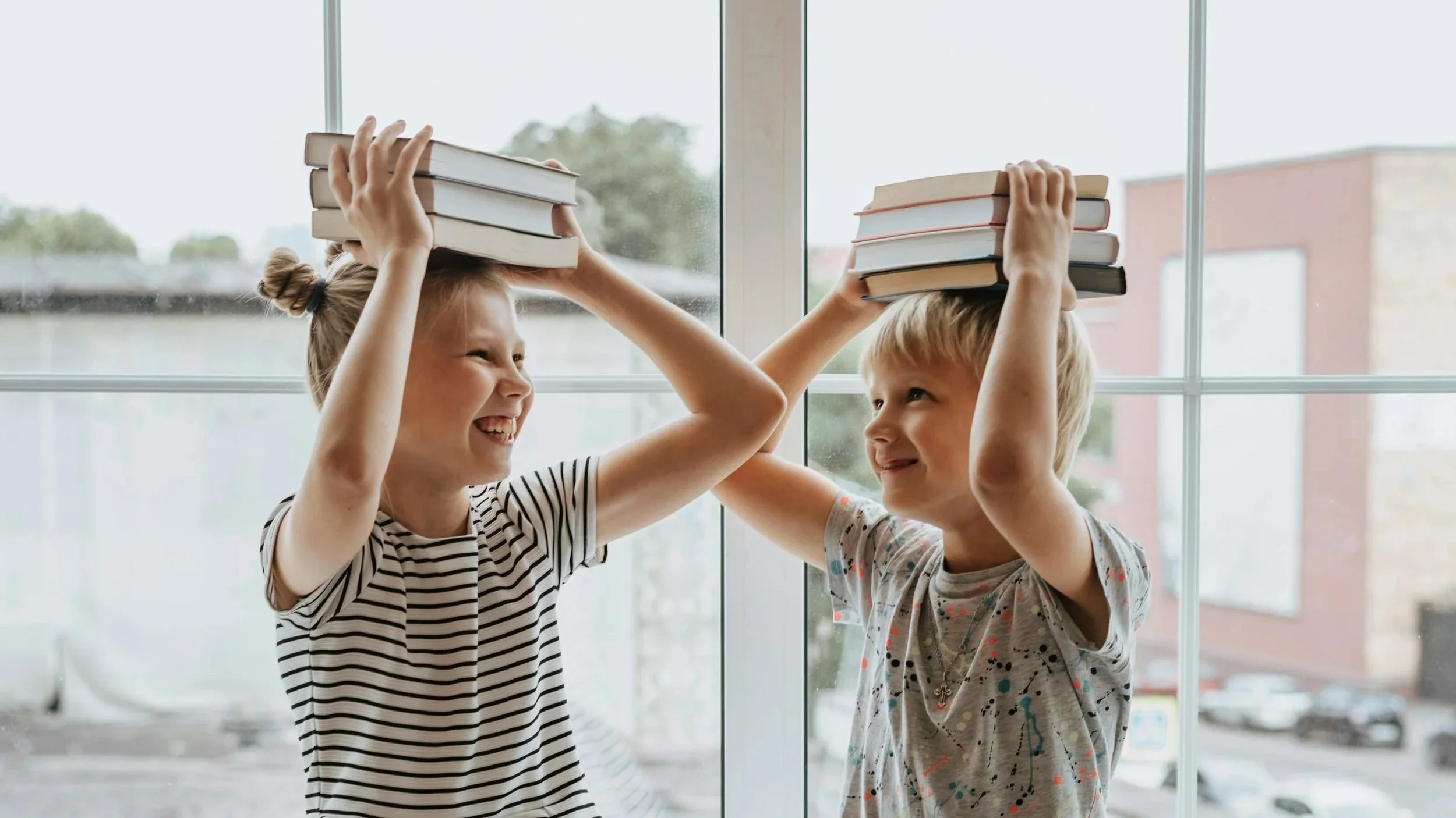 Two children, a girl and a boy, smiling and playfully balancing stacks of books on each other's heads inside near a large window with a city view in the background.