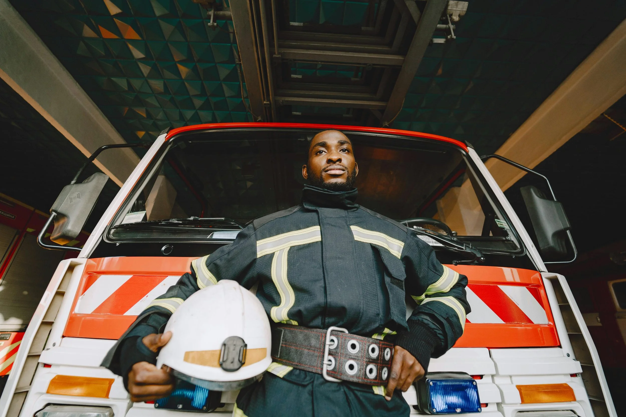 Firefighter standing in front of a fire truck indoors, holding a helmet and wearing a uniform with reflective stripes.