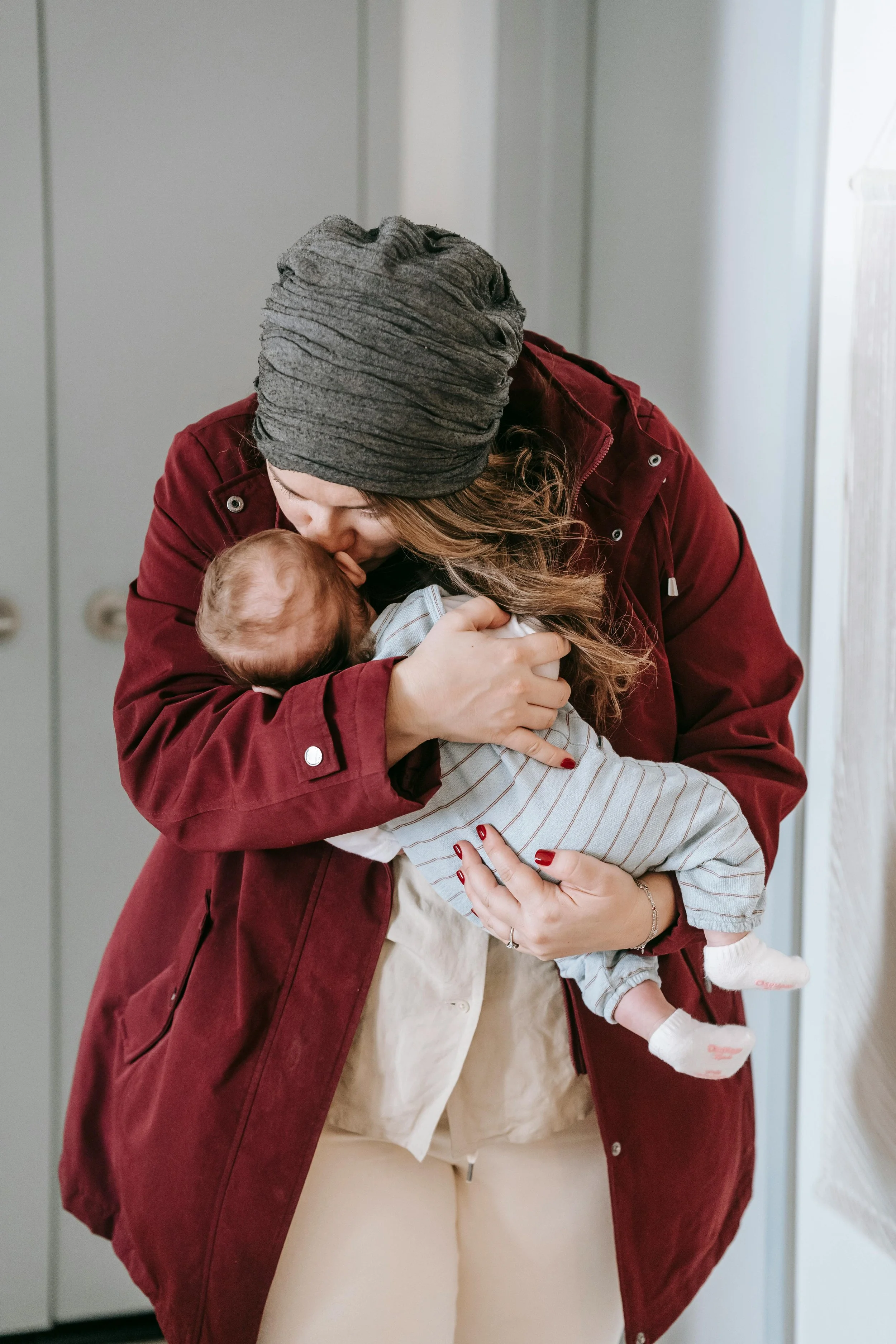 A woman wearing a gray beanie and red coat holding a baby, kissing the baby's head.