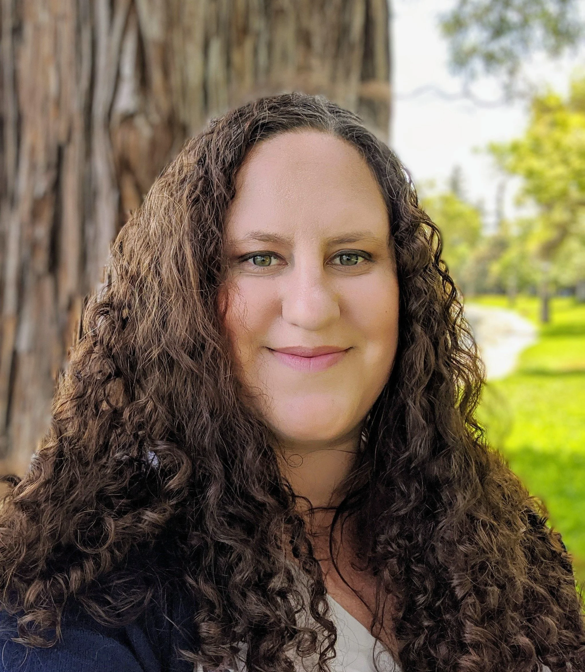 A woman with curly brown hair and hazel eyes smiling outdoors, with trees in the background.