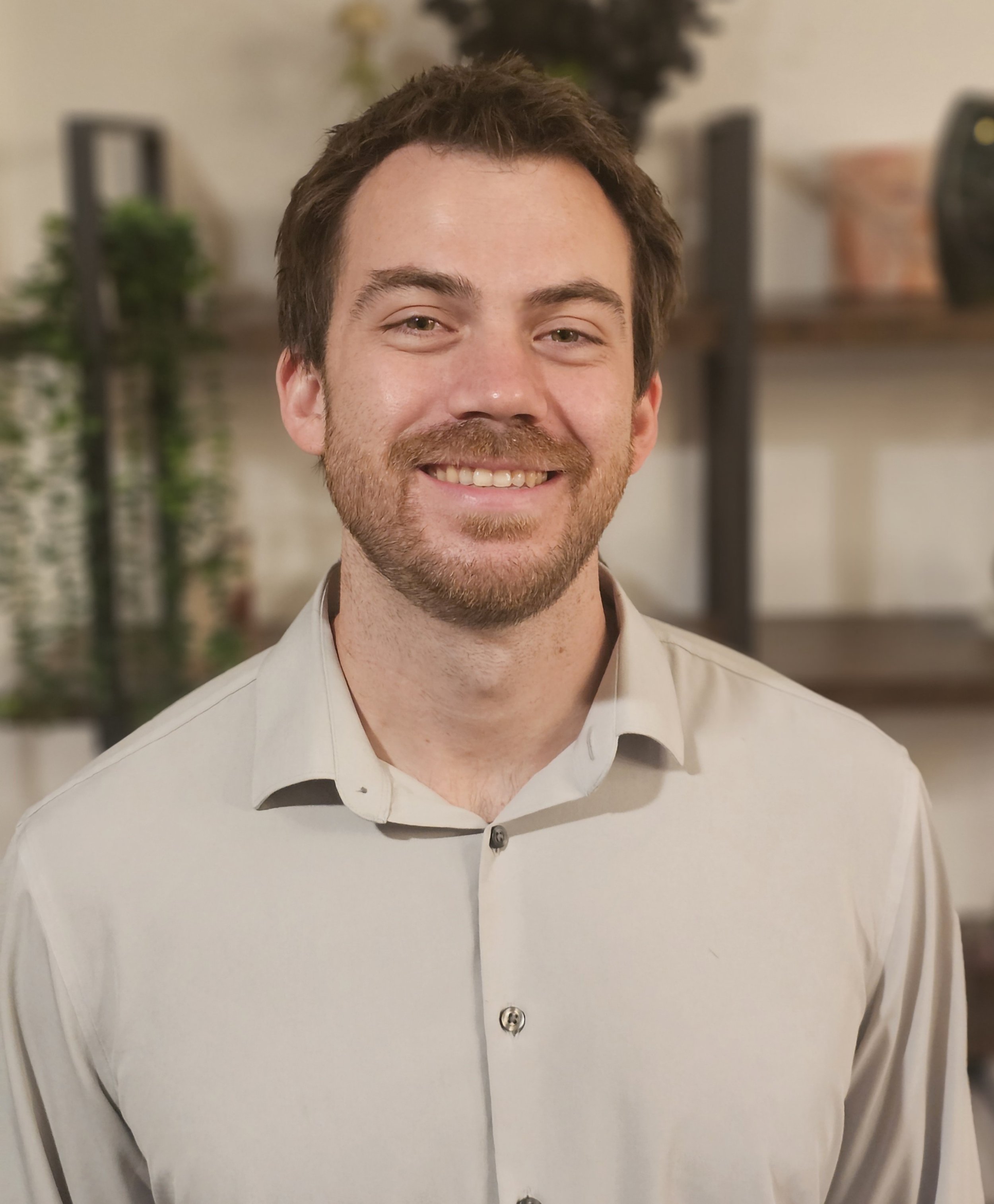 A man with short brown hair, a beard, and light-colored eyes, smiling and wearing a light-colored collared shirt, standing indoors in front of a blurred background of shelves with decorative items and plants.